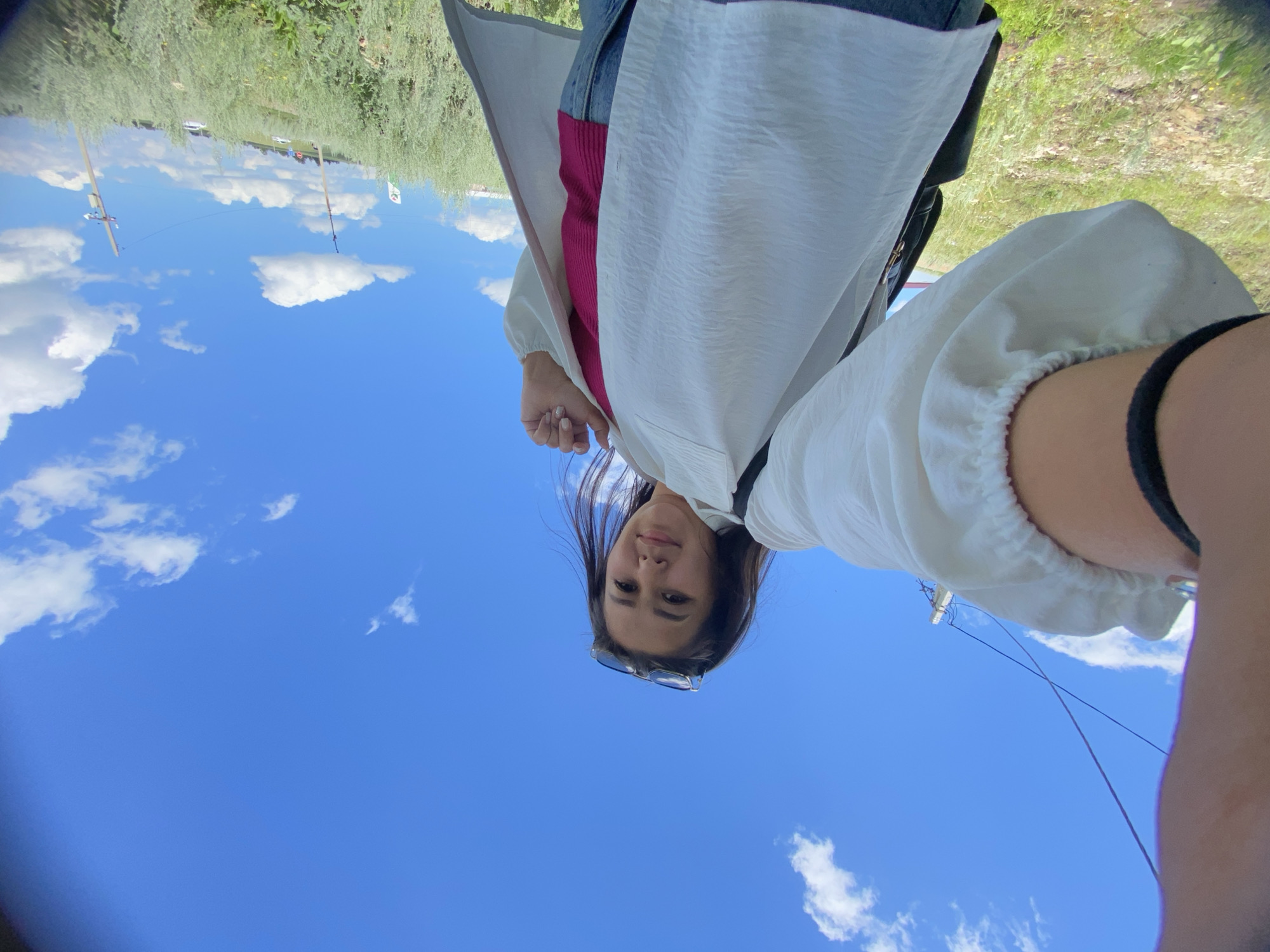 A young woman is taking a selfie outdoors on a sunny day, with a bright blue sky and scattered white clouds above. She is wearing a white open shirt over a pink top and blue jeans. She has sunglasses perched on her head. The background shows a grassy hillside with some bushes and scattered power lines. The overall scene appears to be in a rural or suburban area.