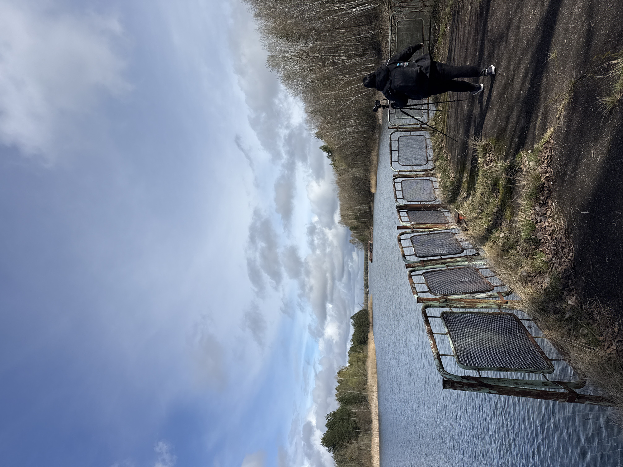 A vertical, outdoor shot depicts a scene in Pripyat, Ukraine. The perspective is from the edge of a water canal, alongside which stands a row of rusted, metal mesh barriers. On the right side of the canal, a paved path runs parallel to the water. A person dressed in black, with a camera mounted on a tripod, stands on the path, presumably taking a photo. On the opposite side of the canal, trees line the bank. The sky is filled with scattered clouds.