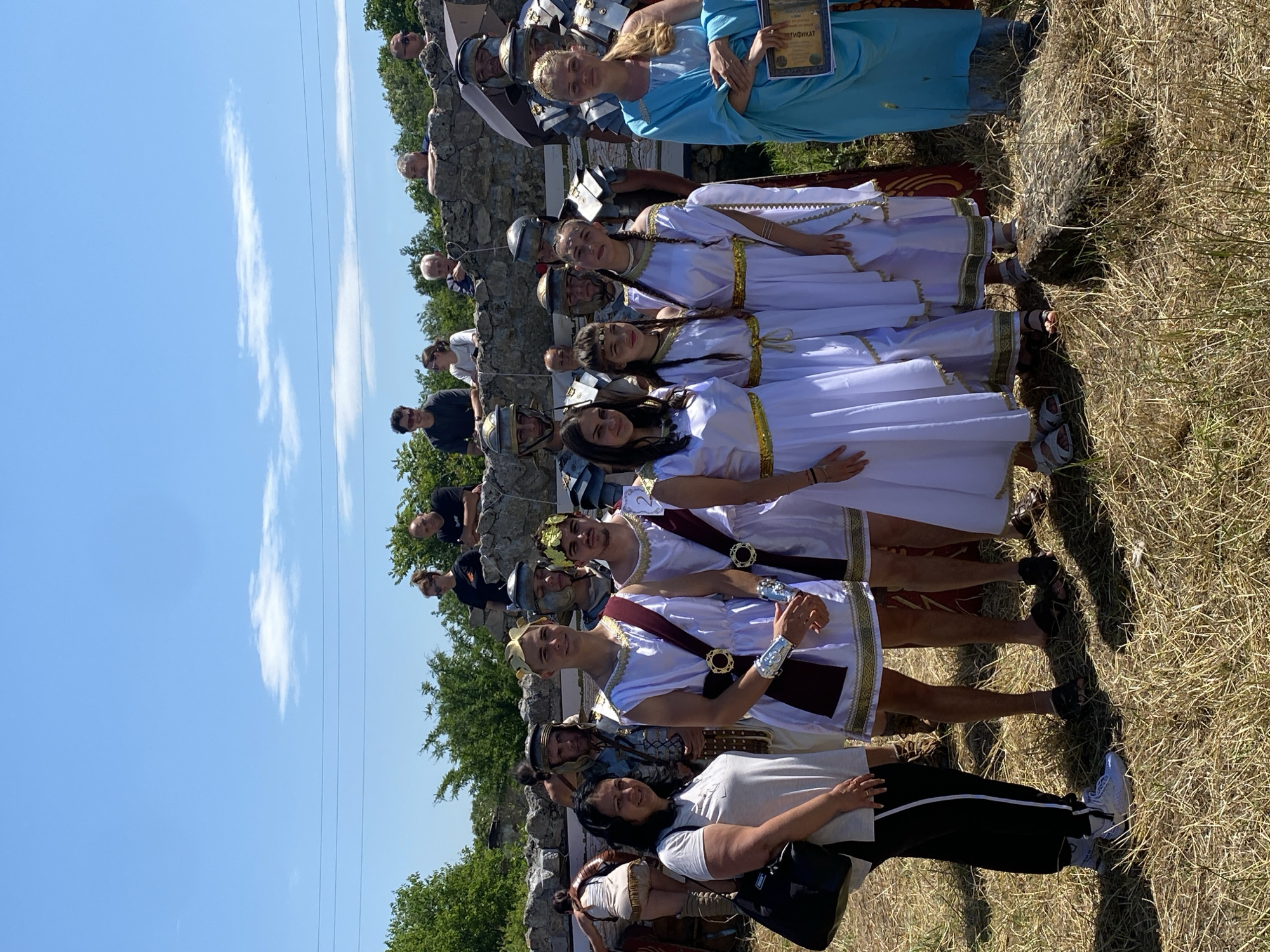 The image depicts a group of twelve individuals standing outdoors on dry, grassy terrain under a clear blue sky in Gigen, Bulgaria.
In the foreground, seven individuals are positioned, primarily facing the camera. From left to right:
1. An adult female with dark hair, wearing a light-colored t-shirt, black pants with white side stripes, and white athletic shoes. She carries a black shoulder bag and smiles at the camera.
2. A young adult male in a white Roman-style tunic with gold trim, a maroon sash, and silver arm cuffs. He wears a gold laurel wreath and smiles at the camera. His feet are covered by dark sandals.
3. Behind the male in a tunic, an adult male is partially visible, wearing Roman legionary armor including a helmet and breastplate. His face is directed towards the camera.
4. A young adult female in a white Roman-style tunic with gold trim, a gold headpiece, and dark braided hair. She smiles slightly at the camera. Her feet are covered by light-colored sandals.
5. A young female in a white Roman-style tunic with gold trim, a gold headpiece, and dark braided hair. She smiles at the camera. Her feet are covered by light-colored sandals.
6. A young adult female in a white Roman-style tunic with gold trim, a gold headpiece, and dark braided hair. She smiles slightly at the camera. Her feet are covered by dark sandals.
7. An adult female with blonde hair, wearing a light blue chiton-style dress. She holds a rolled document in her clasped hands and looks towards the camera.
In the mid-ground, a rough-hewn stone wall extends horizontally behind the primary group. Above and behind this wall, five additional individuals are visible, appearing to observe the scene. They are dressed in modern casual attire. Two power lines traverse the upper left quadrant of the sky. Green foliage and bushes are scattered behind the stone wall. The ground is covered with dry, light-brown grass and sparse vegetation. A dark, rough stone object rests on the ground in the lower right foreground. The overall scene is brightly lit by direct sunlight, casting shadows.
