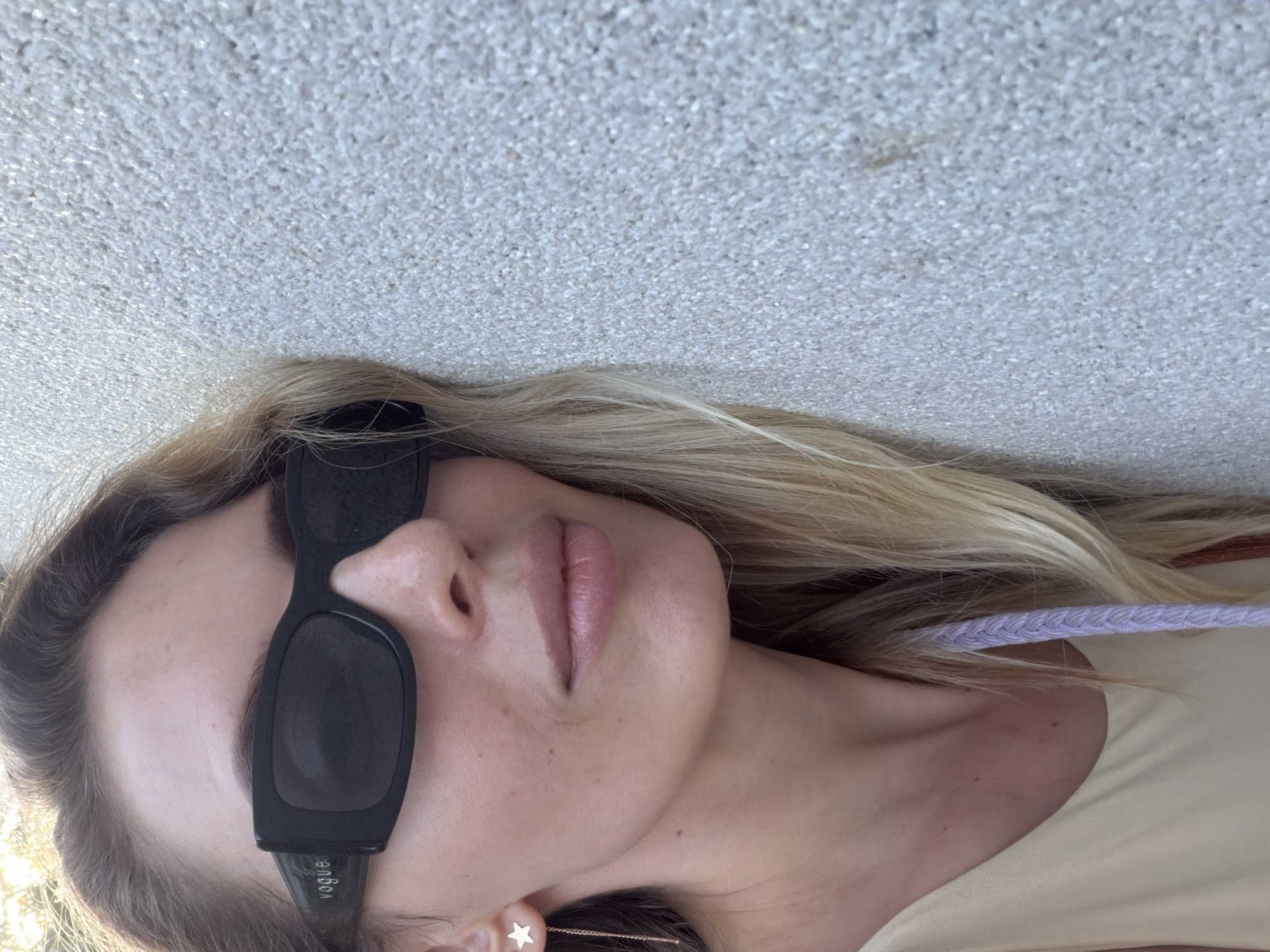 A woman leans her head against a textured, light-colored wall in Budva, Montenegro. She is wearing black sunglasses with the "Vogue" logo visible on the left temple. A star-shaped earring with a dangling gold chain is visible on her left ear. Her blonde hair is long and flows around her face and shoulders. She is wearing a light-colored sleeveless top. A braided purple cord is tucked behind her ear and draped over her shoulder. The sunlight casts a bright glow on her face and hair. The wall appears to be an exterior surface, possibly part of a building or a decorative element.
