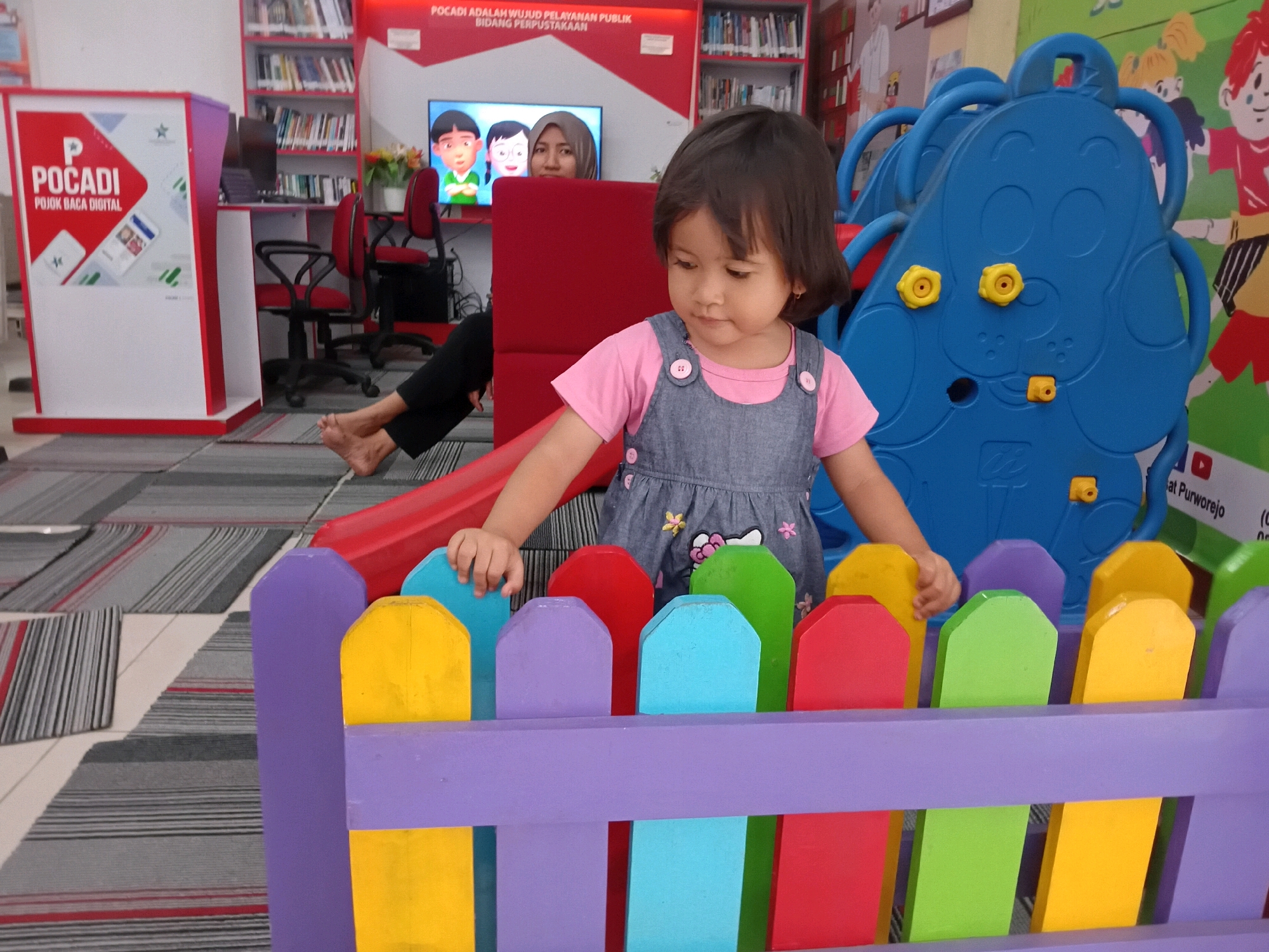 A young girl with dark hair and wearing a pink shirt and a denim overall dress is standing behind a colorful toy fence. She is looking down and her hands are visible touching the fence. Behind her, a woman wearing a hijab is visible, seemingly watching the girl. The scene appears to be indoors, possibly a library or a children's play area, with bookshelves and a television screen displaying cartoon characters in the background. The floor is covered with striped rugs. A sign with text that includes "PURWOREJO" is partially visible in the background, indicating the location.