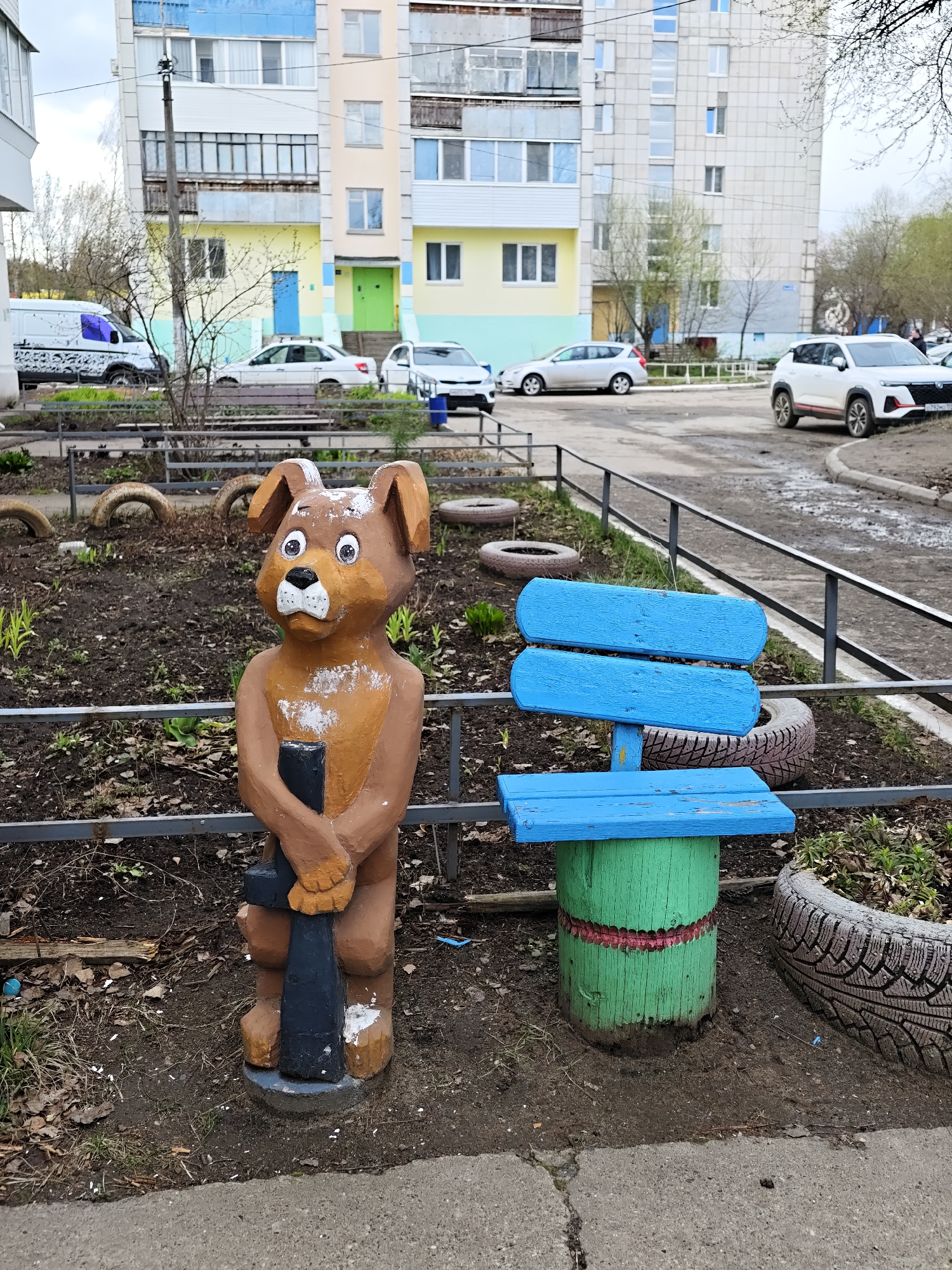 This daytime photograph captures an outdoor scene in a residential area, likely in Chaikovsky, Russia, during early spring. No people are visible.
In the foreground, a brown wooden dog statue stands guard, holding a black object in its paws. The statue has white details around its eyes, nose, and chest, and appears slightly weathered. To its right is a small, child-sized wooden bench, painted bright blue for the backrest and seat, and supported by a green cylindrical base with a red stripe. Both the statue and bench are positioned in a garden bed, partially enclosed by a low metal fence. The ground is dark, muddy soil with sparse, nascent green growth and scattered fallen leaves, suggesting the onset of spring or recent damp weather. Old tires are visible, repurposed as planters or decorative borders.
The midground features an asphalt road or courtyard with several parked cars, including a white commercial van with graphic decals, a white sedan, a silver hatchback, and a white SUV (a partially visible license plate shows "R792MC 159", indicating Perm Krai, Russia). A utility pole stands prominently near the center-left. Beyond the road, several multi-story apartment buildings dominate the background, painted in pastel shades of yellow, light blue, and green, with numerous windows and balconies. Trees and shrubs, mostly bare or just beginning to bud, are scattered throughout the scene.
The sky is overcast, casting a soft, diffuse light over the area. The overall atmosphere is quiet and mundane, typical of an urban residential courtyard. The colorful buildings and whimsical dog statue add touches of visual interest to an otherwise practical setting.