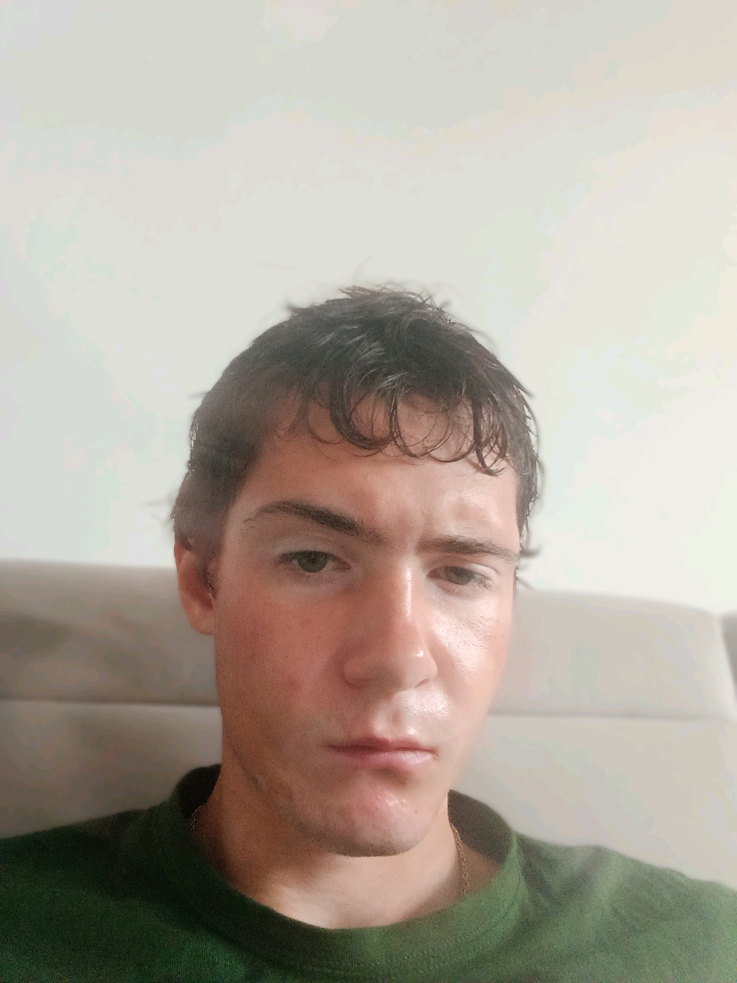 The image is a close-up, waist-up selfie of a young man with dark, tousled hair and fair skin. He is wearing a dark green t-shirt with a thin gold chain visible around his neck. The background is a plain, light-colored wall with a soft, neutral-colored headrest or couch behind him. The lighting is bright and diffused, creating a slightly washed-out effect. The man's expression is neutral, with his eyes looking slightly downward and to the side. The image does not contain any information that allows for its placement in Szczecin, Poland, or any specific city.