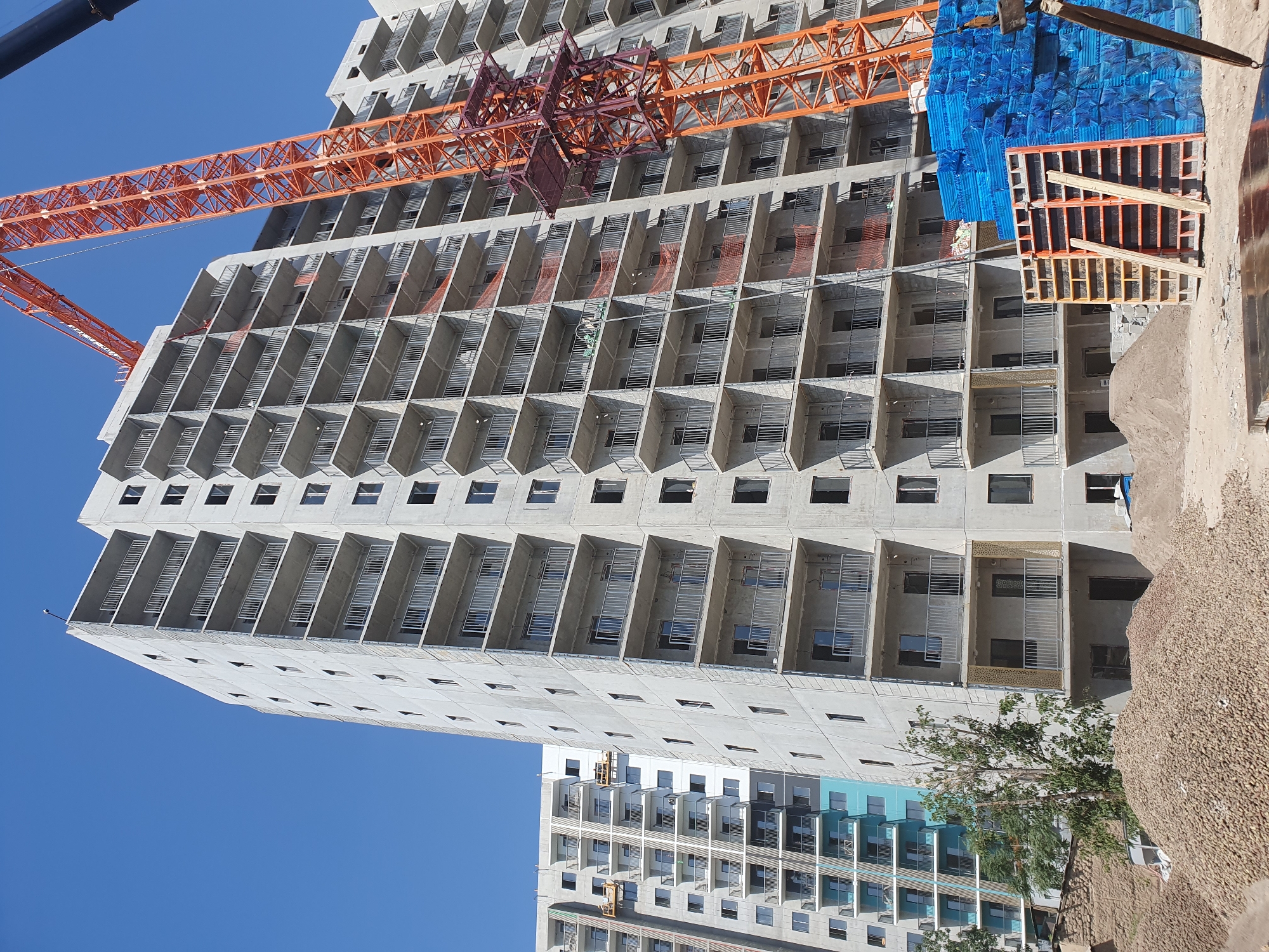In Tashkent, Uzbekistan, a multi-story residential building is under construction under a clear blue sky. The building is made of concrete with numerous balconies, each equipped with metal railings. An orange construction crane with a red support structure is positioned next to the building. In the foreground, piles of gravel and sand are visible, along with stacks of blue-wrapped construction materials and orange formwork. The scene suggests active construction work is in progress.