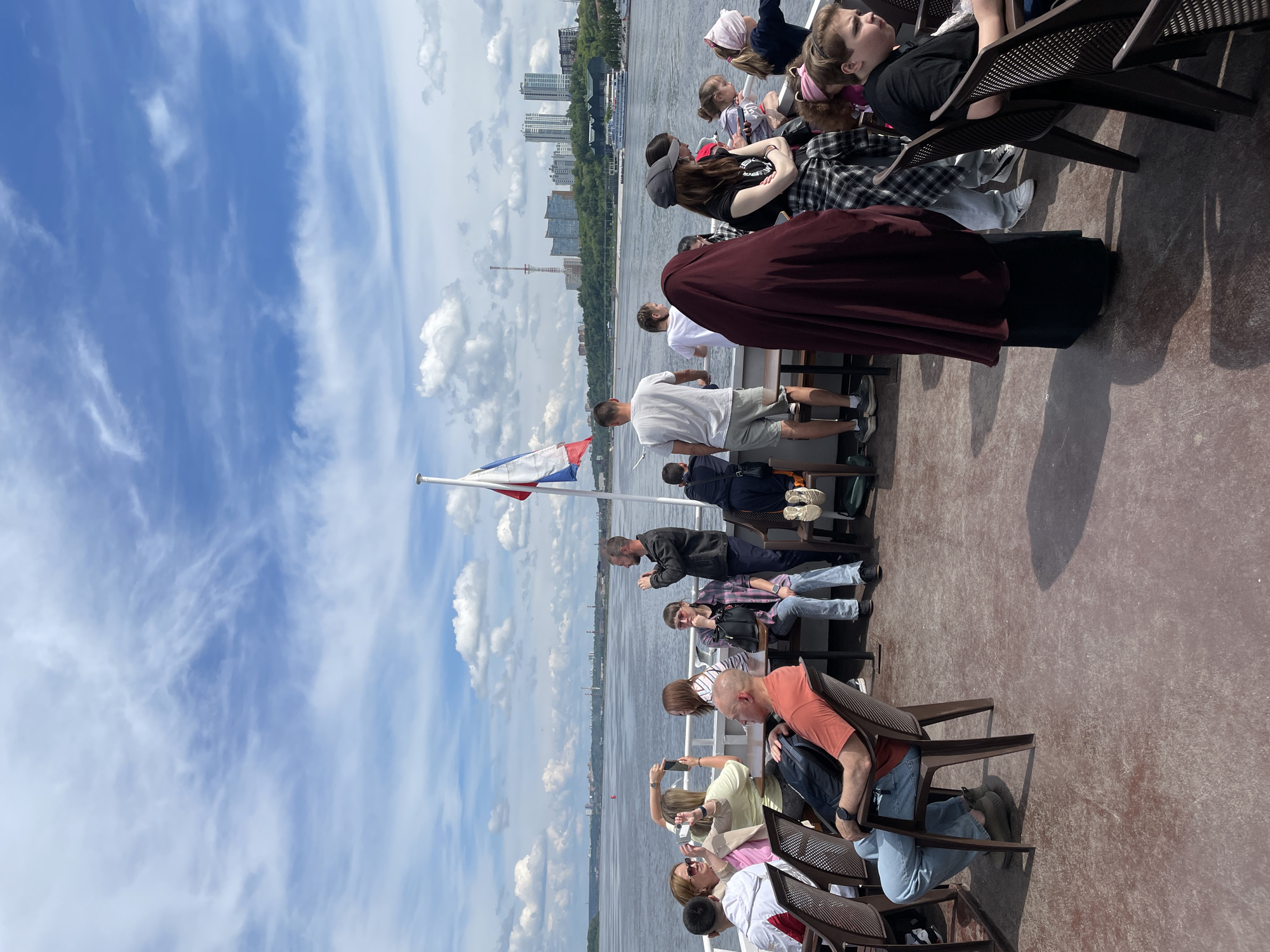 This image captures a vibrant daytime scene aboard a passenger boat, likely cruising along the Kama River in Perm, Russia. The perspective is from the deck, looking out over the water towards the city skyline.
Numerous people are visible, seated in brown, plastic-like chairs arranged in rows. They appear to be passengers enjoying a scenic tour. Most individuals are casually dressed in t-shirts, shorts, and light clothing, suitable for warm weather. Many are looking out at the expansive river and the distant shore, suggesting they are taking in the views. Some are conversing, while one person in the foreground is holding a smartphone, seemingly capturing the surroundings. A distinct figure in a dark maroon hooded garment stands prominently among the seated passengers.
The far bank of the river showcases a mix of modern high-rise buildings nestled among green trees, under a bright blue sky dotted with scattered white clouds, indicating pleasant weather. A tall, thin industrial structure or communication tower is also visible on the distant horizon. At the bow of the boat, a flagpole proudly flies the Russian flag, a horizontal tricolor of white, blue, and red. The sun casts noticeable shadows on the boat's deck, confirming it is daytime, likely late morning or afternoon. The overall atmosphere appears relaxed and pleasant. There is no visible text in the image other than the flag design.