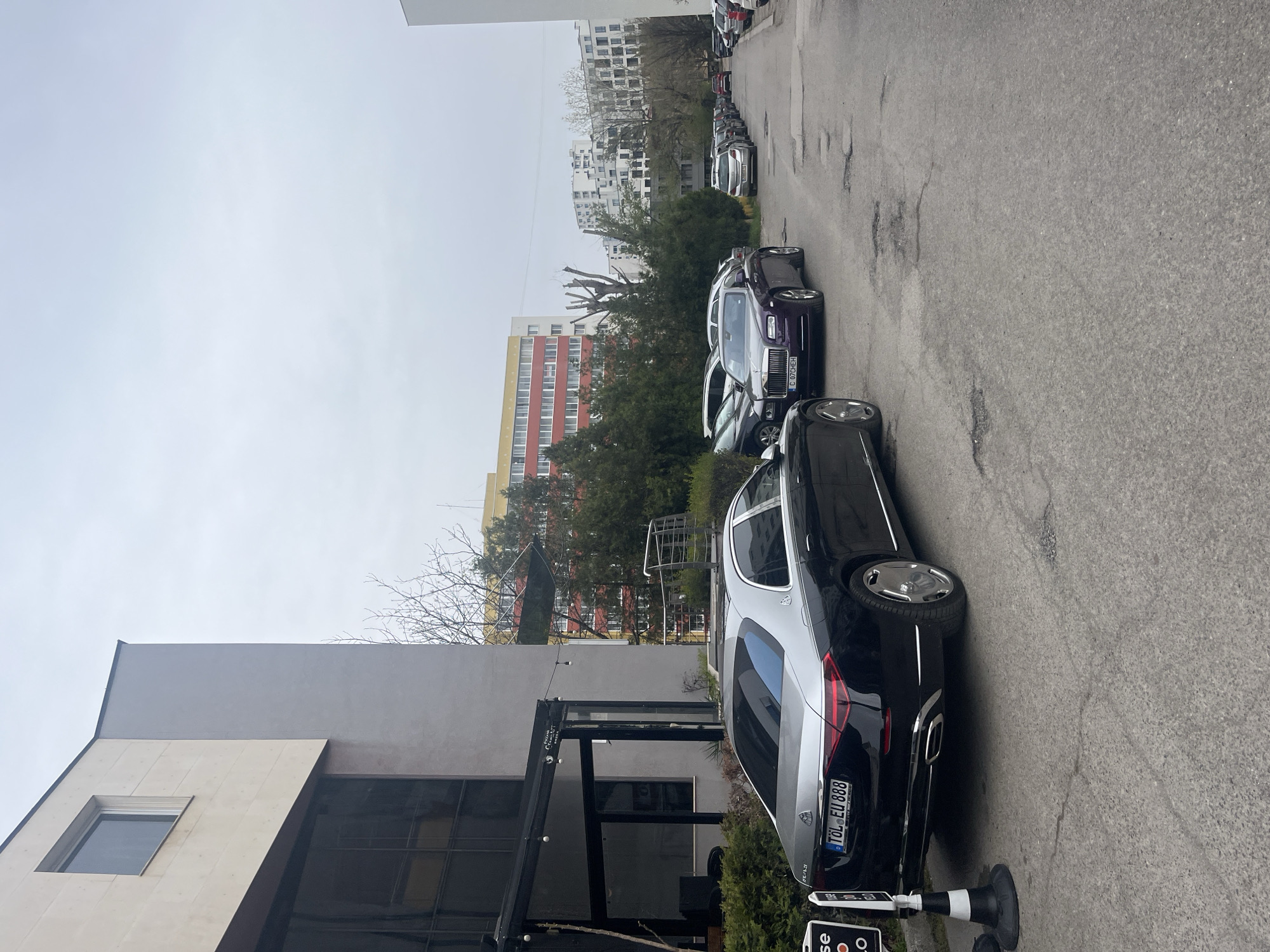 The image depicts a street scene in Sofia, Bulgaria, characterized by a line of parked cars. In the foreground, a black and silver Mercedes-Maybach with license plate "TÖL EU 888" is parked next to a building with a modern architectural style. The building features a large glass facade and a retractable awning. Adjacent to the Maybach, a dark purple Rolls Royce is also parked. Further down the street, numerous other cars are parked along the narrow, cracked asphalt roadway. In the background, there are apartment buildings with varied color schemes and bare trees against an overcast sky. The general atmosphere suggests a residential or commercial area, possibly in a central part of the city. No people are visible in the image.