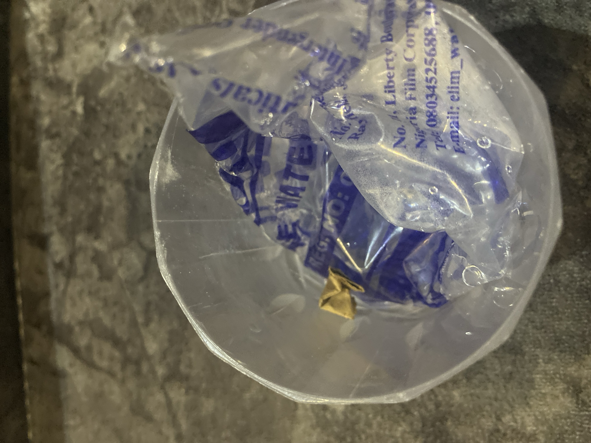A clear plastic cup contains a crumpled piece of paper with blue lettering on it, possibly a label or packaging. The cup is placed on a textured, dark surface, which appears to be a countertop or table. The background is out of focus and shows a mottled, greyish-brown pattern. The label inside the cup has text that is partially visible, including words like "WATER", "REG", "No. 4. Liberty Bod", "Nigeria Film Corps", "Tel: 08034525688", and "E-mail: elim_wa". The overall lighting suggests an indoor environment. No specific location in Jos, Nigeria is discernible from the image.