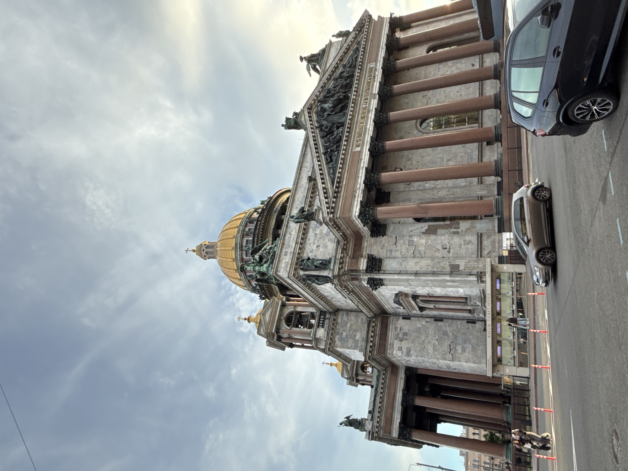 This is a low-angle, wide shot of Saint Isaac's Cathedral in Saint Petersburg, Russia, on a cloudy day. The grand cathedral, with its imposing marble facade and golden dome, dominates the frame. The sky above is a mix of blue and gray clouds, with the sun breaking through on the right side of the image, casting a warm glow.
In the foreground, a street is visible with two cars, a silver hatchback and a black SUV, driving on it. The hatchback is parked to the left of the SUV. The street is bordered by a sidewalk with a fence, and behind the fence, a ticket office with the text "КАССЫ - TICKET OFFICE" is visible. A few people are standing near the ticket office, and a small group of individuals is walking on the sidewalk to the left of the ticket office.
The cathedral's architecture is intricate, featuring large columns, sculptures, and ornate detailing. The golden dome gleams in the sunlight. The overall impression is one of grandeur and historical significance. The image captures a moment in time, with the daily life of the city unfolding against the backdrop of this magnificent landmark.