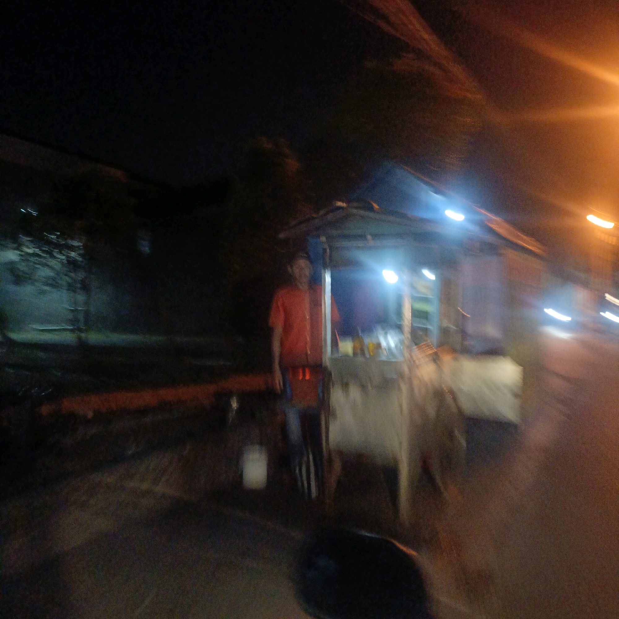 The image captures a street scene at night in Salatiga, Indonesia, likely near a roadside stall. A person, appearing to be a vendor, is standing in front of a small, open-air structure that serves as a food stall. The vendor is wearing a red t-shirt and what appears to be an apron. The stall is illuminated by a bright light, possibly from a bulb, and there are items on display, including what could be bottles or containers. The background is dark, with hints of trees or buildings and blurry streaks of light, suggesting movement or ambient city lighting. The overall image is somewhat blurred, indicating it was taken with a moving camera or in low light conditions. The ground in front of the stall appears to be paved.