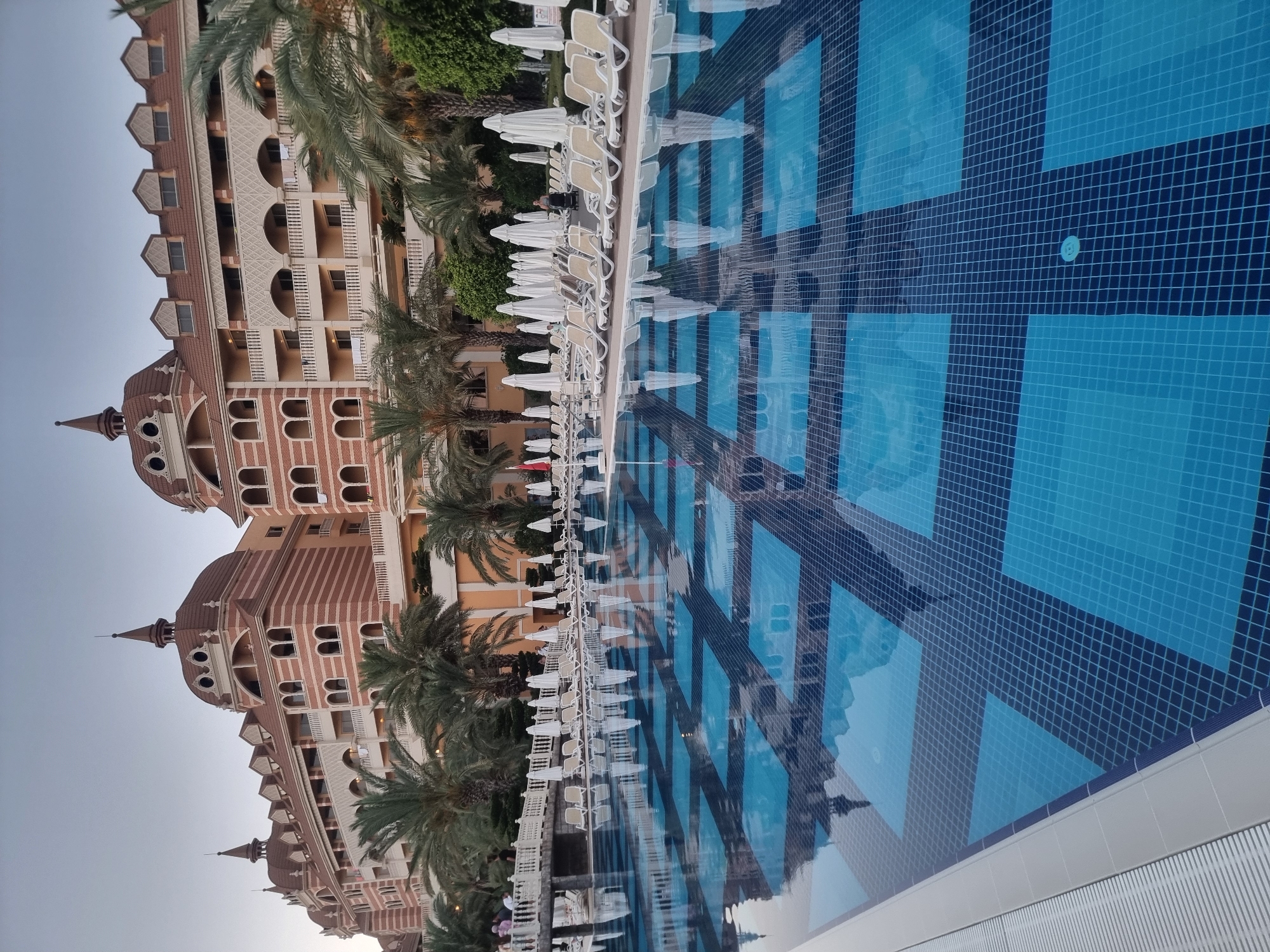 The image captures a serene resort scene in Manavgat, Turkey, featuring a large swimming pool in the foreground and a grand hotel complex in the background. The pool's surface reflects the architecture and palm trees, creating a mesmerizing pattern with its blue tiled squares. Deck chairs and white umbrellas are neatly arranged around the pool, suggesting a leisurely atmosphere. Several people are visible near the pool and on the hotel's balconies, engaged in various activities. The hotel's distinctive architecture, with its red brick facade, ornate domes, and arched windows, speaks to a Mediterranean or Ottoman influence. The overall impression is one of a tranquil and luxurious vacation destination.