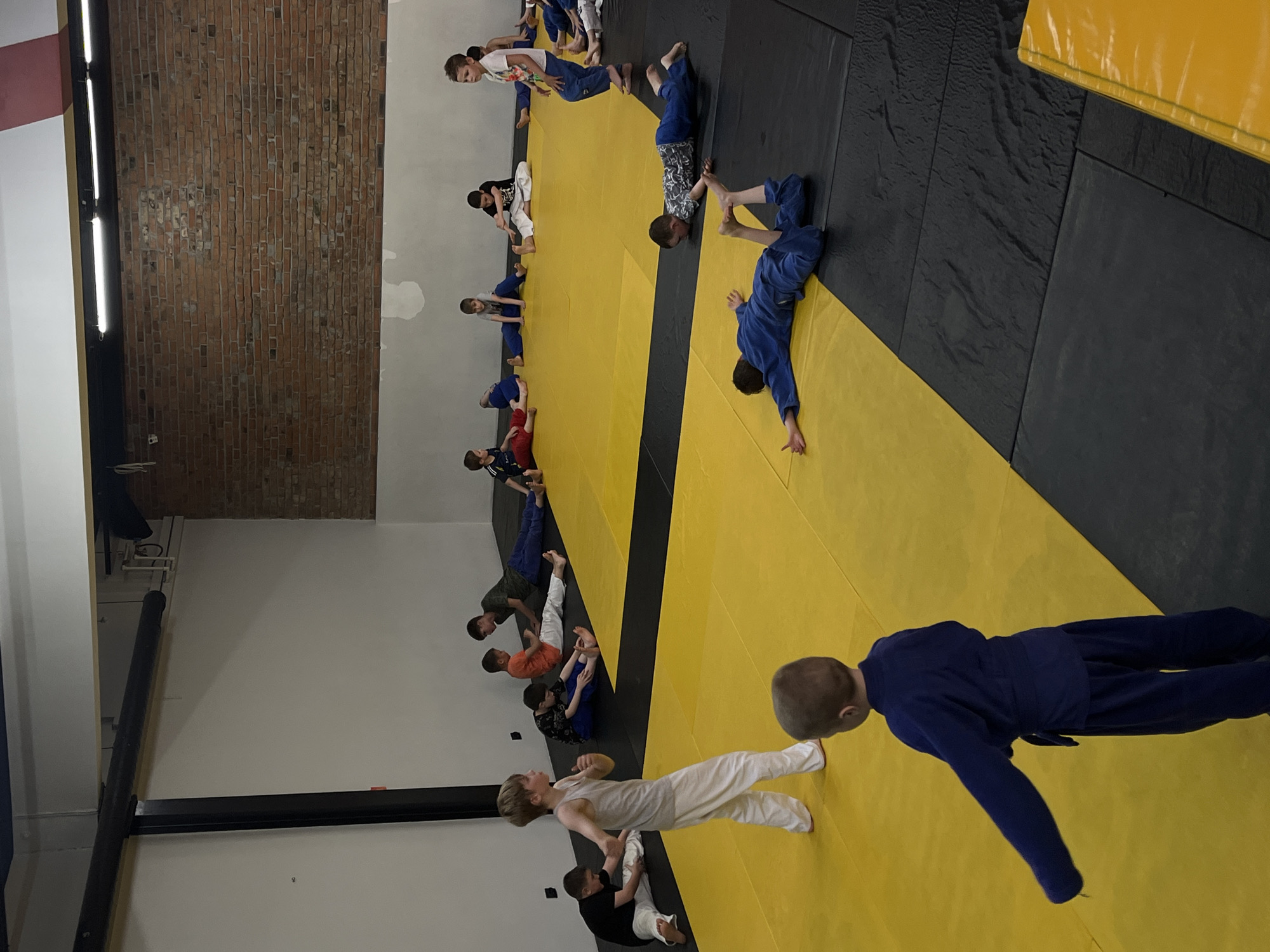 In a martial arts studio in Krasnoyarsk, Russia, a group of young boys are engaged in a judo class. The studio features a large mat area with alternating yellow and black sections. The walls are painted white, with a brick wall visible in the background. Several boys are in various stages of a warm-up or stretching exercise, some sitting on the mat with their legs extended, while others are lying down. One boy, wearing a light grey tank top and white pants, stands in the foreground, facing away from the camera, appearing to be demonstrating a movement. Another boy in a dark blue judogi is also in the foreground, facing away from the camera. The overall atmosphere suggests a training session for young athletes.