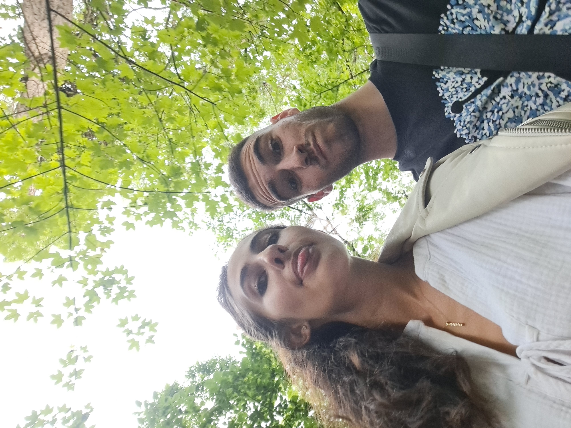 An outdoor selfie shows a young woman and a young man in Lokorsko, Bulgaria, with lush green foliage and a bright sky in the background. The woman, positioned on the left, has dark, curly hair and is sticking her tongue out slightly. She is wearing a white textured shirt with a tie-front and a light-colored jacket. A delicate gold necklace is visible around her neck. The man, on the right, has short brown hair and a beard. He is wearing a black t-shirt with a blue and white abstract pattern. Both individuals are looking towards the camera, and the perspective suggests they are looking up. The scene appears to be set in a natural, possibly forested area.