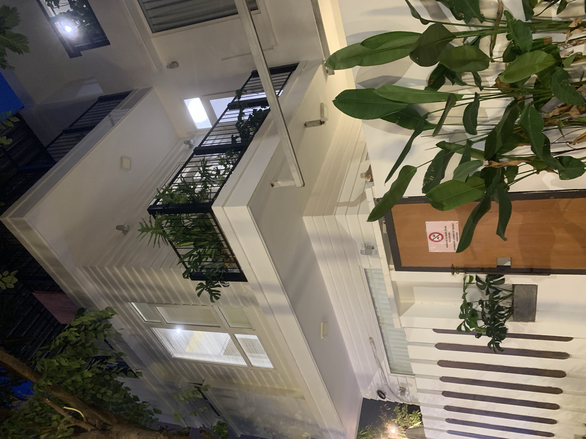 The image depicts the exterior of a modern multi-story building at night, likely a residential or commercial property, located in Đà Nẵng, Vietnam. The building features clean white facades with horizontal siding, and several balconies with black railings. Large windows with blinds are visible on the upper floors, with interior lights illuminating the rooms. Lush green plants, including large-leafed tropical varieties, are placed on the balconies and in planters near the ground level. A wooden door is visible on the right side of the image, with a sign featuring a red circle with a diagonal line through it and text in what appears to be Vietnamese and English, stating "NO PARKING IN FRONT OF THE EXIT THANK YOU!". The foreground is dominated by large green plants and a white fence with dark vertical stripes. The overall scene suggests a well-maintained property with a tropical aesthetic, characteristic of coastal cities like Đà Nẵng. There are no people visible in the image.