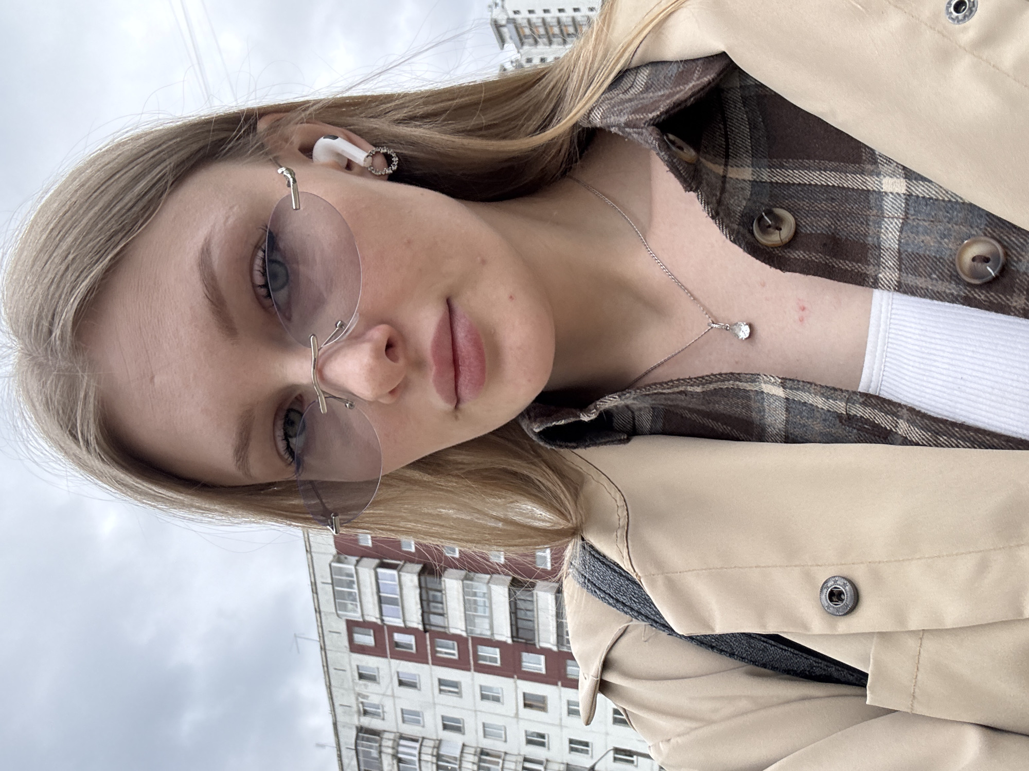 A young, fair-skinned woman with blonde hair and blue eyes is depicted in a selfie. She's wearing frameless round sunglasses and a white wireless earbud in her left ear, accessorized with a silver necklace. She's dressed in a plaid shirt underneath a light beige trench coat.
In the background, a multi-story residential building is visible under an overcast sky. Based on the architectural style of the building and the coloring, this image appears to be taken in Perm, Russia.