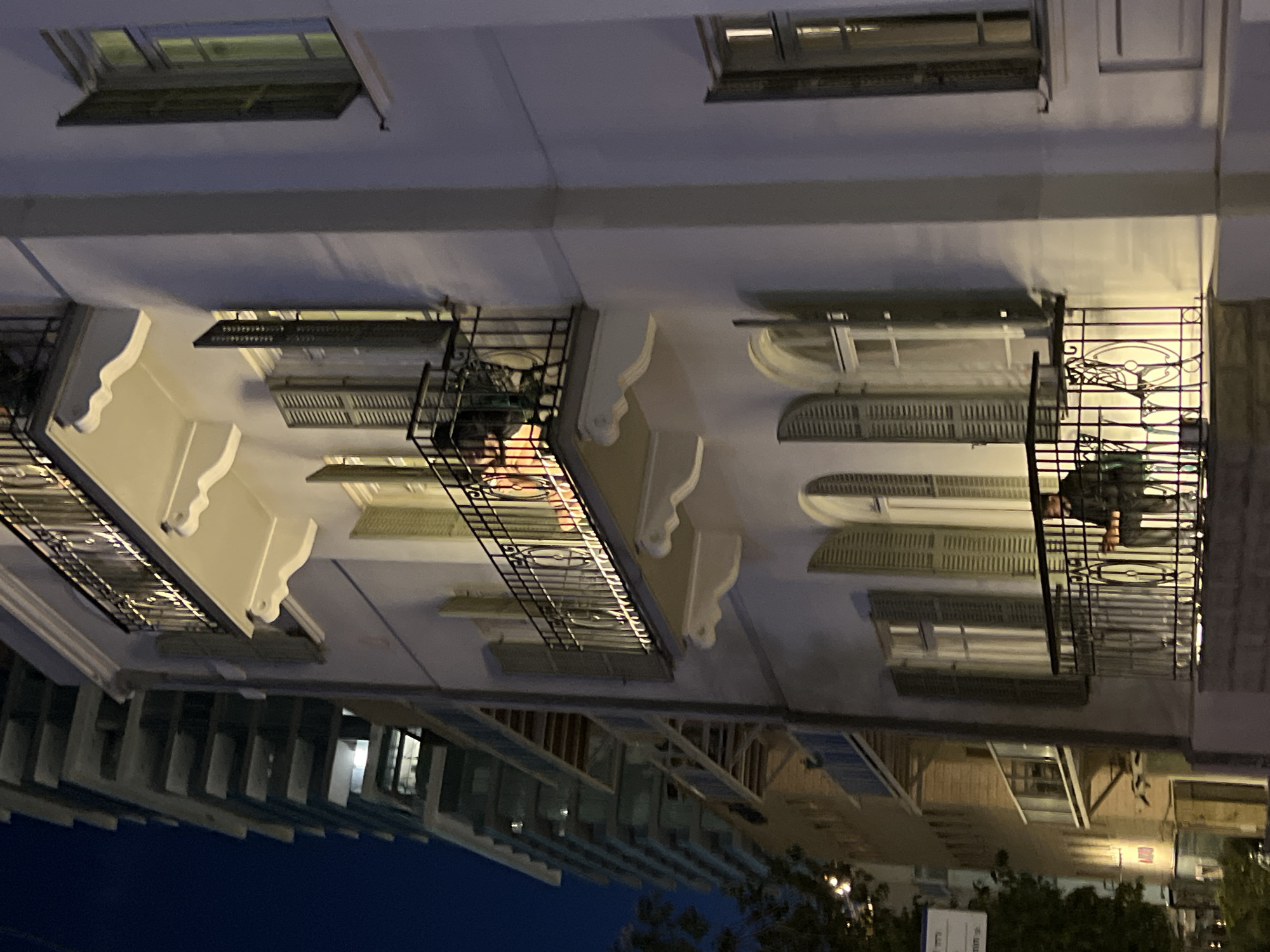 The image shows a low-angle view of an apartment building in Tel-Aviv, Israel, at night. The building is white with ornate architectural details, including decorative balconies. Some balconies have wrought iron railings. Windows are visible with closed shutters. A person is standing on one balcony. The lower portion of the image shows additional buildings and some trees. Streetlights are visible, indicating an urban environment. The sky is dark blue.