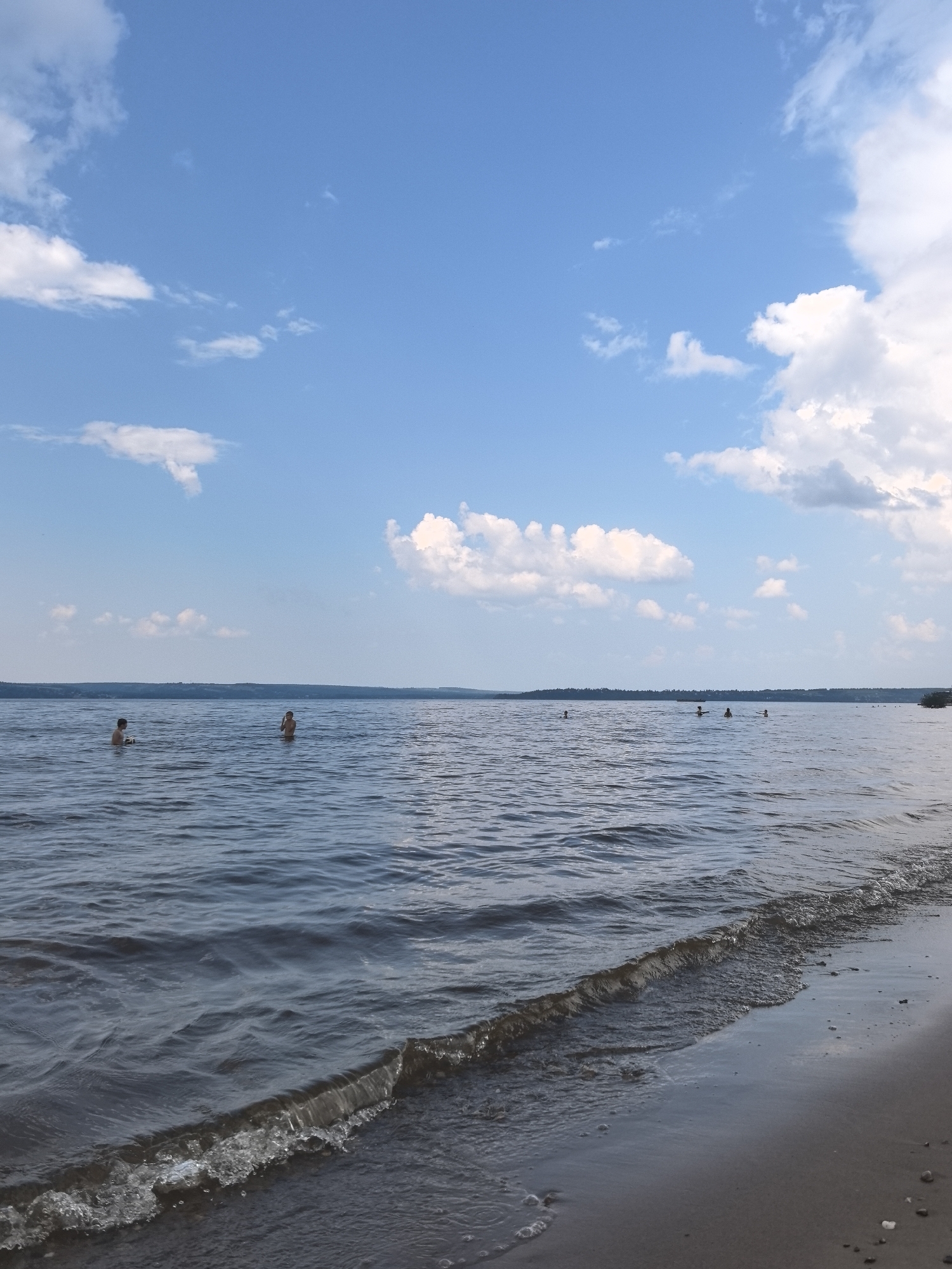 The image depicts a natural landscape scene with a body of water and a sky with clouds. In the foreground, a sandy shore is visible, with a gentle wave lapping at the edge. The water extends into the middle ground and background, appearing calm with ripples. Several individuals are in the water at varying distances from the shore. The sky is predominantly blue with scattered white clouds. The horizon shows a distant treeline. While the image itself does not provide specific locational information within Perm, Russia, it presents a common depiction of a natural recreational area that might be found near a body of water such as the Kama River or one of the city's lakes.
