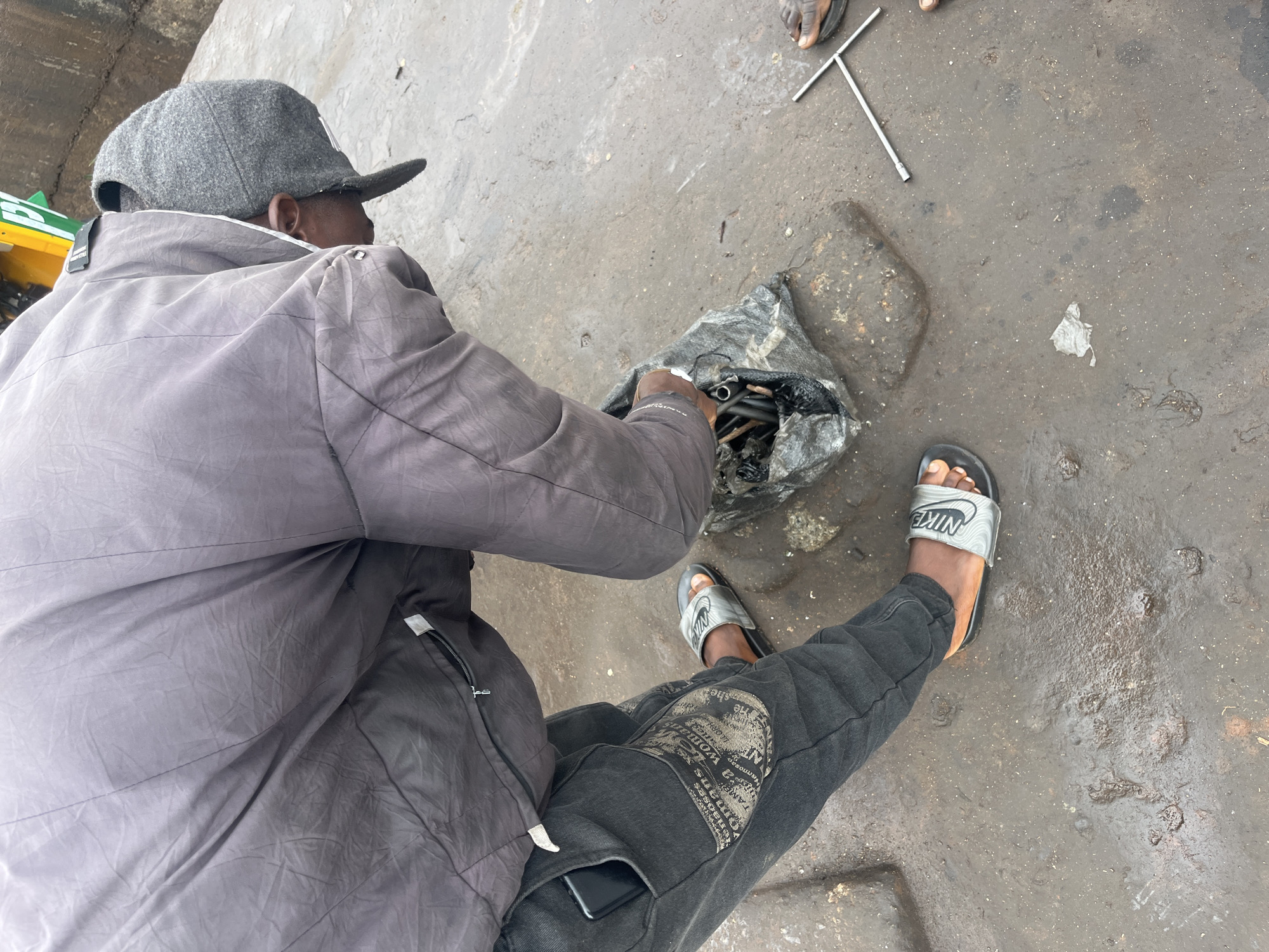 In Bukuru, Nigeria, a person wearing a grey jacket, black jeans, and black and silver Nike slides is leaning over a black plastic bag filled with mechanical parts. The person is wearing a grey cap and appears to be sorting through the contents of the bag. The ground is wet and appears to be a paved or concrete surface. A metal T-shaped tool is visible on the ground to the right of the bag. The overall scene suggests a person engaged in mechanical work or repair in an outdoor environment.