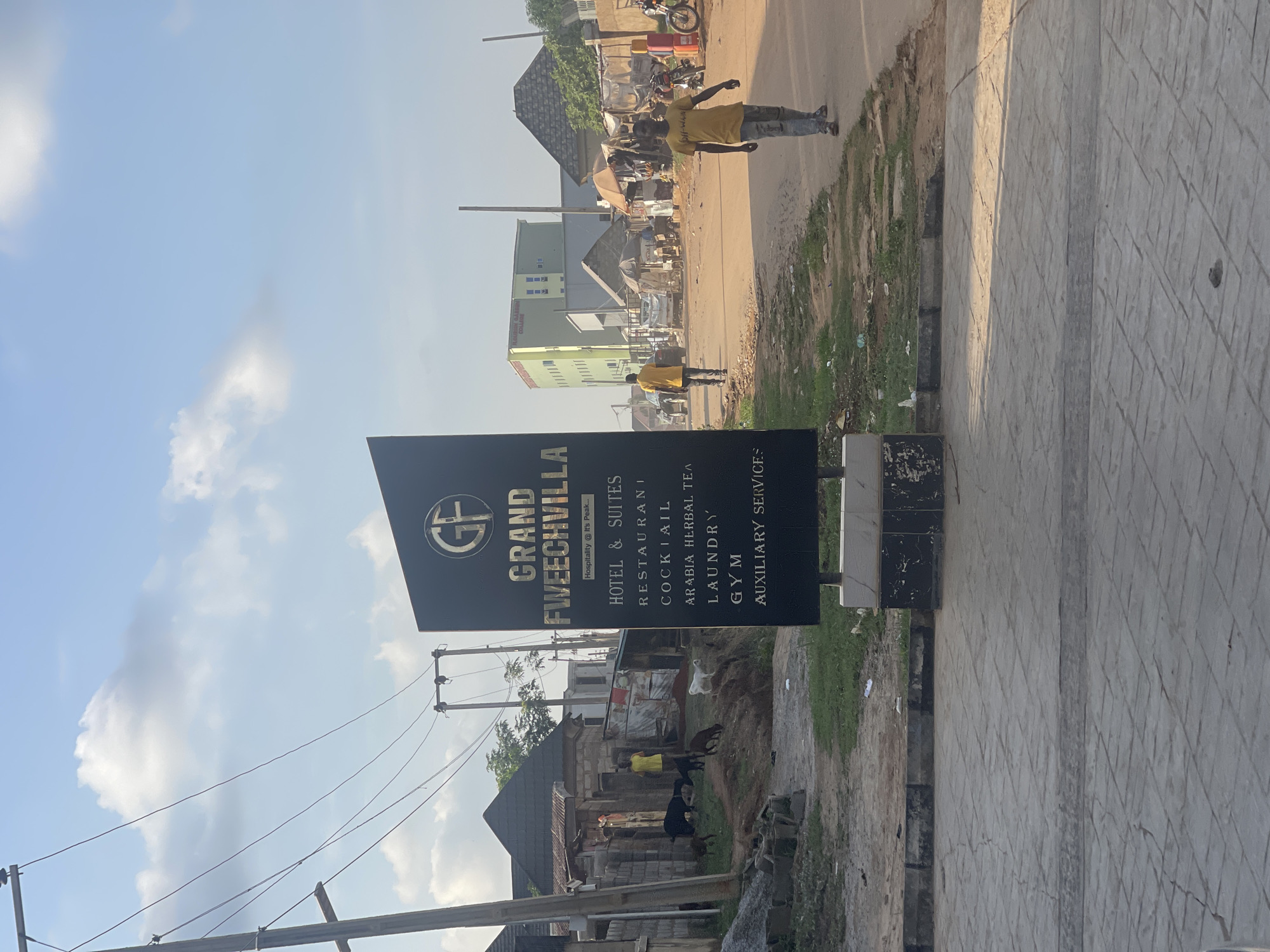 The image depicts a street scene in Abuja, Nigeria, featuring a sign for "Grand Fweechvilla" hotel and suites. The sign, colored black with gold lettering, lists amenities such as a restaurant, cocktail bar, herbal tea, laundry, gym, and auxiliary services. A person in a yellow shirt walks along the roadside in the distance, and a second person is further in the distance wearing a yellow shirt. The road appears unpaved and bordered by vegetation on one side and a tiled sidewalk on the other. Behind the sign, a cluster of buildings and homes suggests a residential or commercial area. Power lines are visible in the sky.