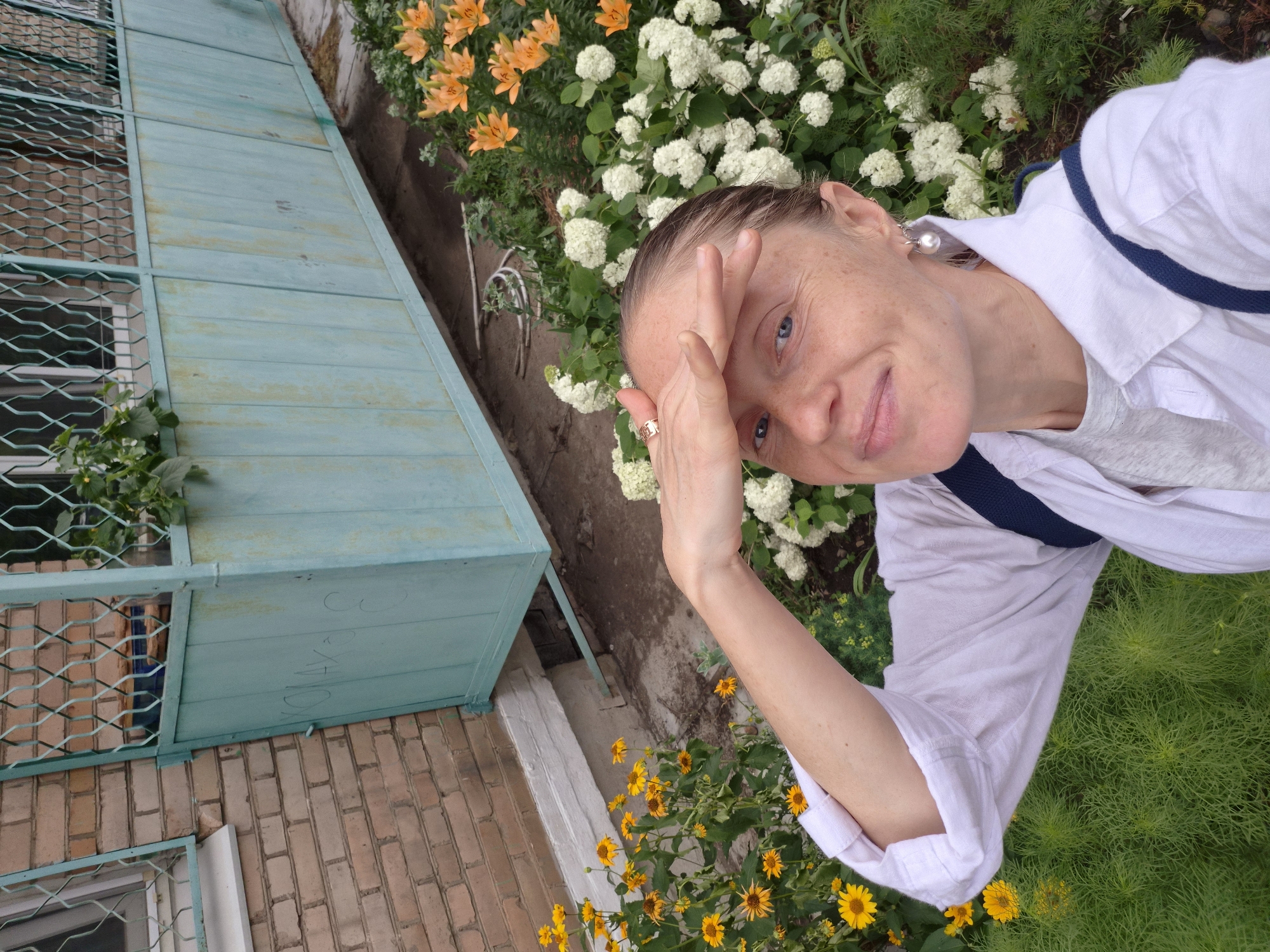 In this close-up, high-angle, self-portrait photograph, a fair-skinned woman with light blue eyes and freckles is positioned in the foreground. She is wearing a white button-down shirt over a grey t-shirt, and a dark blue strap is visible over her shoulder, suggesting she is wearing a backpack. Her right hand is raised, shielding her eyes from the sun, and she is looking directly at the camera with a slight smile. She wears a gold ring on her right index finger and a pearl earring in her left ear.
The background reveals an outdoor setting with lush greenery and blooming flowers. To the left, yellow flowers are in bloom, and to the right, clusters of white hydrangeas and orange lilies are visible. Behind the flowers, a turquoise metal structure, possibly a balcony or fence, is present. The structure is weathered and shows signs of graffiti. To the left of this structure is a brick building.
The image is situated in Arseniev, Russia, as indicated by a portion of the graffiti on the turquoise structure that spells out "ОЛАНЧ С". The overall scene depicts a casual moment outdoors, likely in a residential area given the proximity of the building and garden.