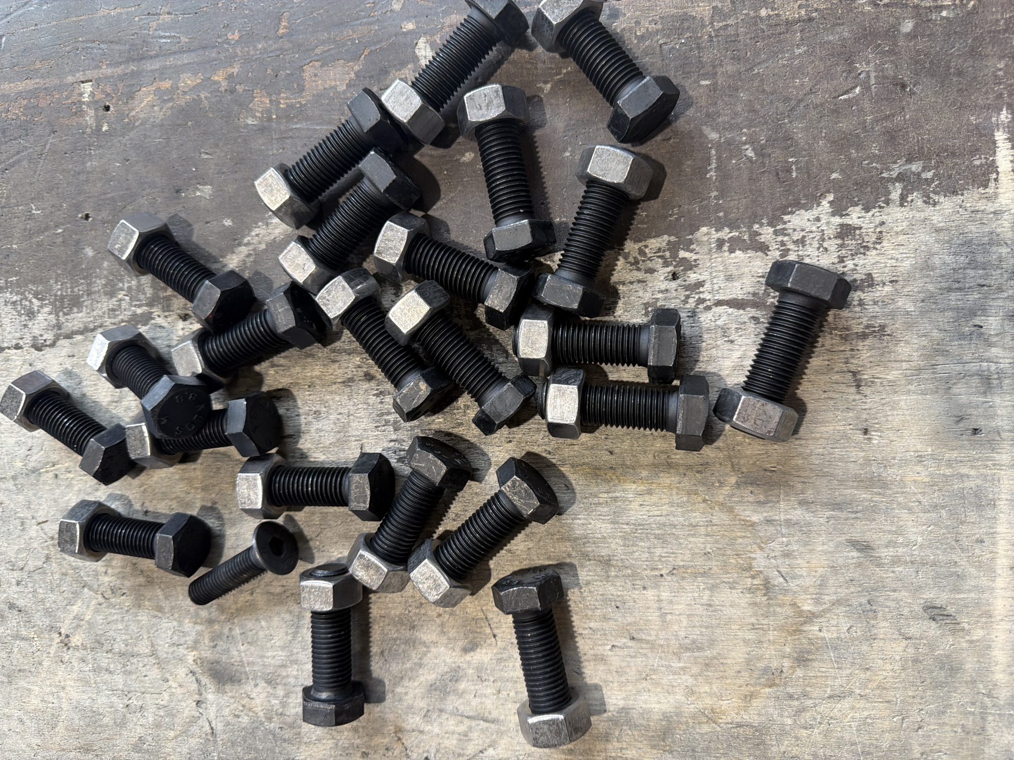 The image displays a collection of hexagonal head bolts and one Allen head bolt scattered across a weathered, textured wooden surface. The bolts are primarily black with silver-colored hexagonal nuts on many of them. The wood surface appears to be a workbench or floor, with visible grain, scratches, and what may be oil stains or a patina of age. The lighting suggests it is daytime, with shadows cast by the bolts indicating an overhead light source. The scene is a close-up, top-down view of these hardware items. There are no people present. The arrangement of the bolts appears random, as if they have been dropped or are waiting to be used. The overall impression is one of a workshop or maintenance area. There is no discernible text visible in the image.