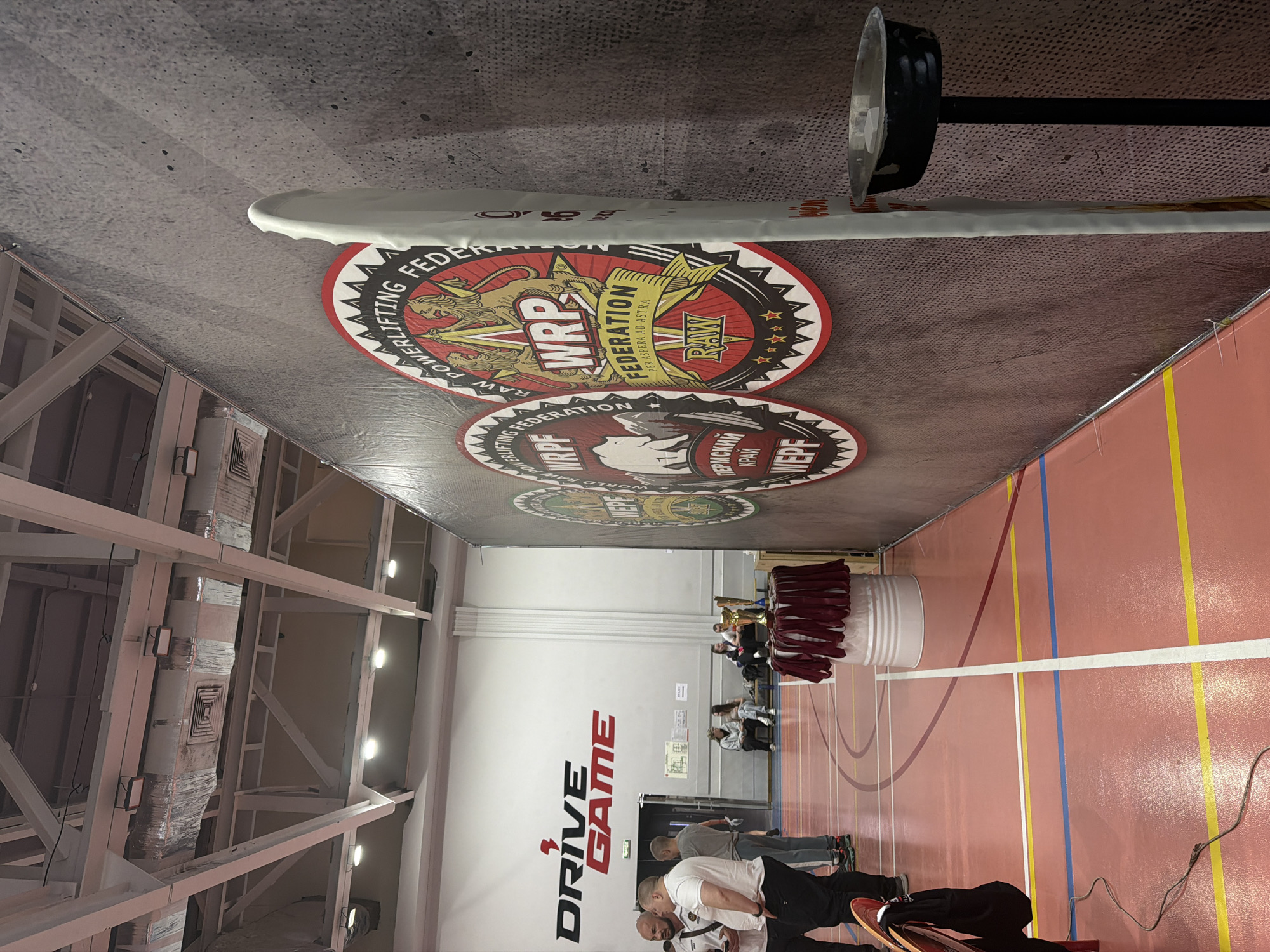 The image shows the interior of a large indoor arena, likely a gymnasium or sports hall, in Perm, Russia, as suggested by the presence of signage in Russian Cyrillic script. The perspective is tilted upwards, focusing on the ceiling area.
A black and silver barbell sits in the upper right, a white-covered weight on the lower left.
Several round banners depicting the "World RAW Powerlifting Federation (WRPF)" logo are attached to the ceiling. The banners are red, black, and white, and include an image of a lion or similar creature.
Below the banners, along the wall, is the word "DRIVE" in large black letters, with "GAME" below and to the right.
In the background, people are visible, some seated and others standing. There's a white cylindrical object draped with maroon cloth, possibly a table or platform, with trophies on top.
The floor is marked with colored lines, and structural beams and lighting fixtures are visible on the left side of the image. The walls are white.