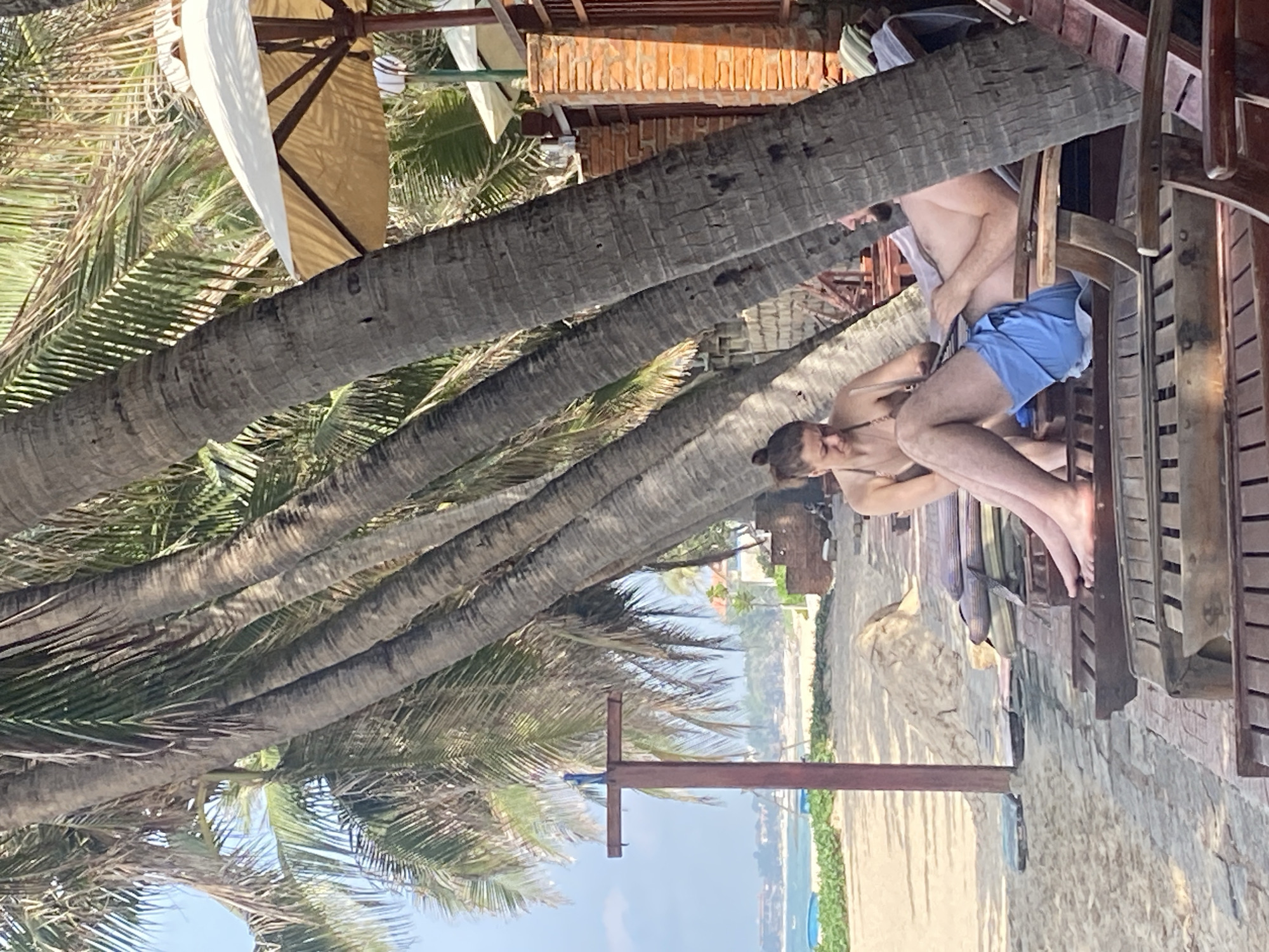 A couple relaxes on lounge chairs under palm trees on a beach in Phu Thuy Ward, Vietnam. The woman, with her hair in a bun, wears a bikini and appears to be applying sunscreen to her legs. The man, wearing blue swim trunks, reclines on a lounge chair, his hand resting on his leg. A beach, ocean, and some buildings are visible in the background. The scene is lit by sunlight, creating shadows from the palm trees.