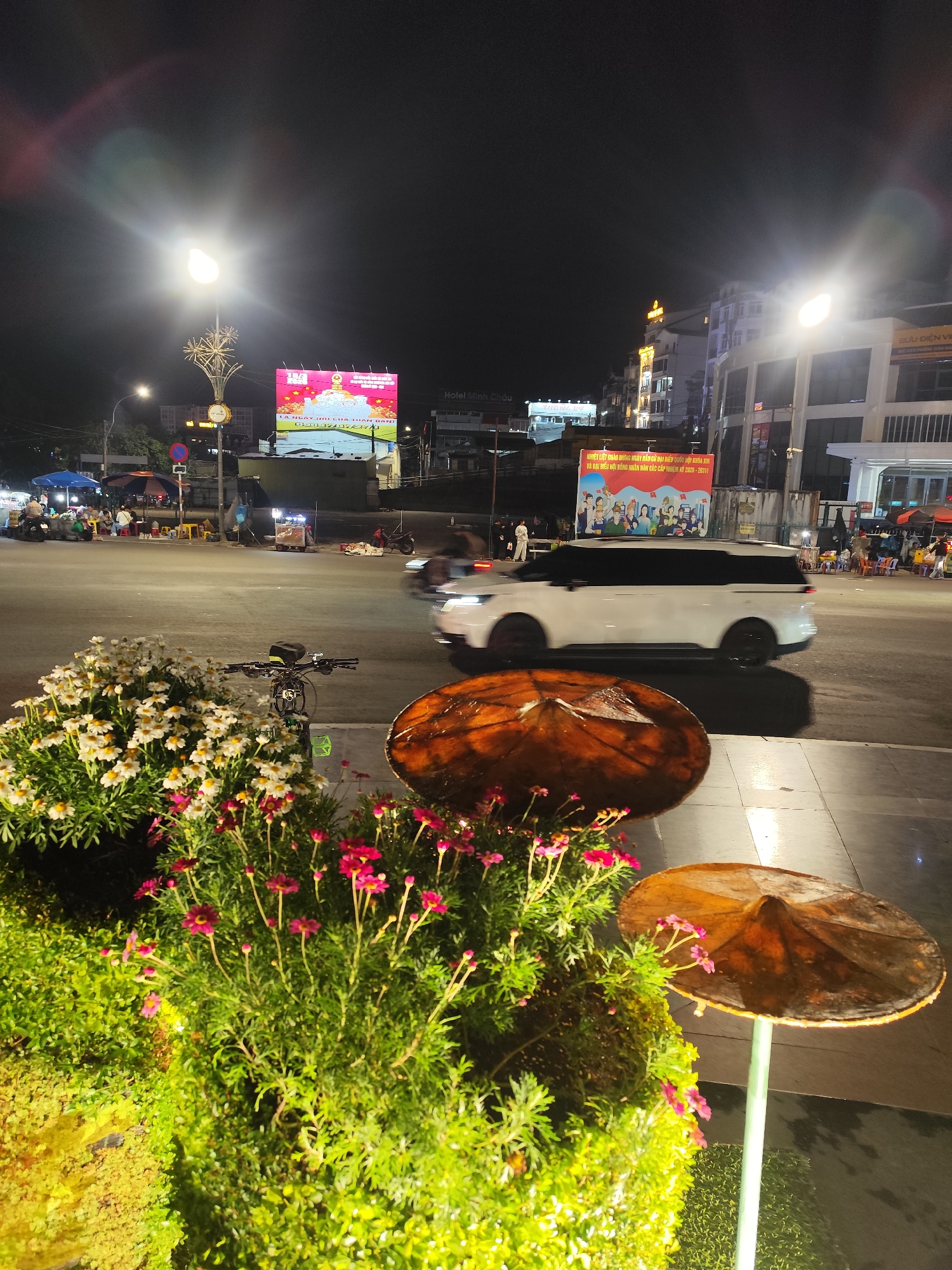 The image depicts a street scene at night in Da Lat, Vietnam. The foreground features a planter with white daisies and pink flowers, next to street decorations mimicking traditional Vietnamese conical hats. Behind this, a white minivan crosses an intersection. Beyond the intersection, illuminated billboards and building facades are visible. People are gathered at small street food stalls and seating areas along the road. Streetlights illuminate the scene. A bicycle is parked near the planter.