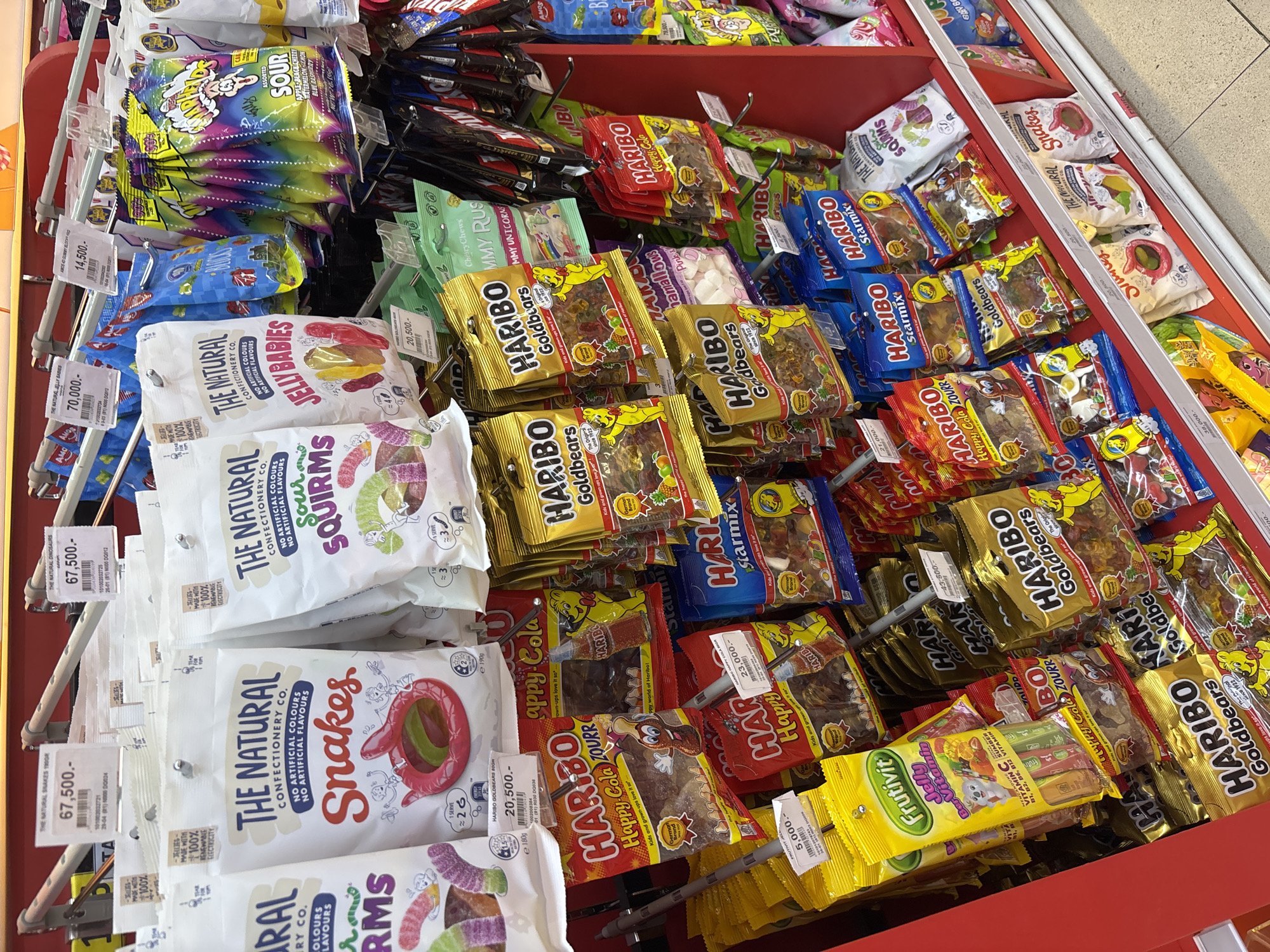 A high-angle, medium-shot photograph depicts a shelf in a retail setting, likely a supermarket or convenience store, filled with various types of candy. The candy is arranged in a densely packed, haphazard fashion, with different brands and varieties mixed together. Prominent brands include Haribo, known for its Goldbears and Happy Cola varieties, and "The Natural Confectionery Co.," featuring products like "Snakes," "Sour Squirms," and "Jelly Babies." Other candies include "Frutivit" and a rainbow-colored "Waw" sour candy. Price tags are visible, indicating the cost of each item, with prices such as 67,500, 70,000, 20,500, 23,000, 5,000, and 14,500, possibly Indonesian Rupiah (IDR). The shelf itself is constructed from red metal, and a portion of the floor can be seen. There are no people visible in the shot. Given the pricing on tags, the brand names, the product types, and the language and format of the price tags, this image was most likely taken in Kuta Selatan, Indonesia.