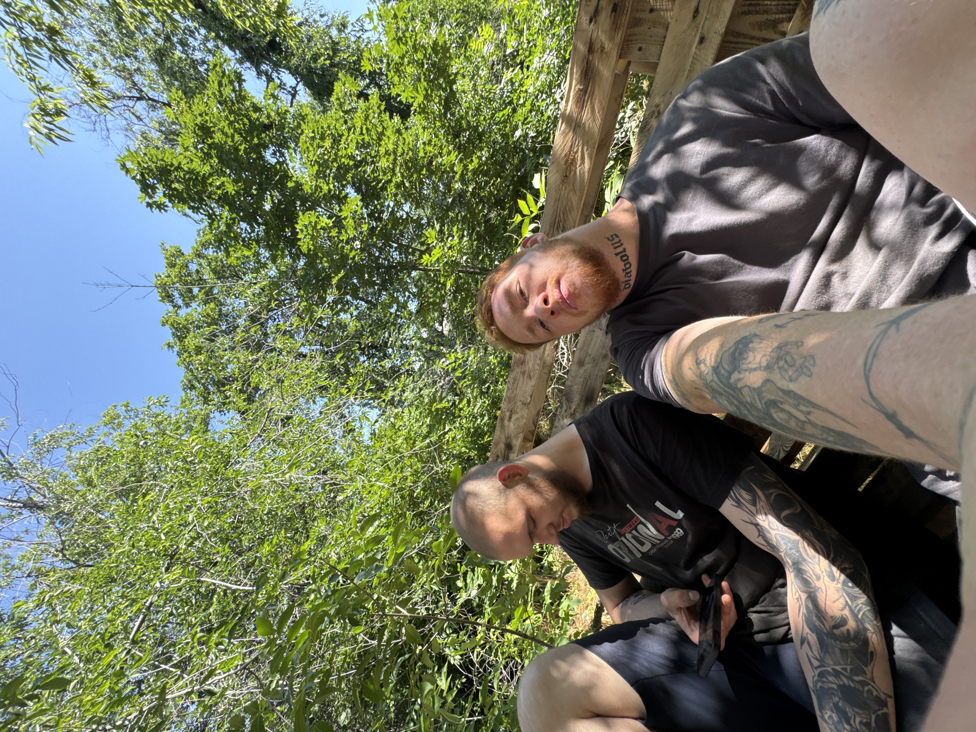 Two men are seated on a wooden bench outdoors. The man on the left has a shaved head and is wearing a black t-shirt. He is looking down at a smartphone in his hand, his left arm covered in intricate tattoos. The man on the right has red hair and a beard. He is wearing a dark grey t-shirt and has a tattoo on his neck that reads "diabolus". His right arm is also heavily tattooed. Both men are positioned in the foreground, with lush green foliage and a bright blue sky visible in the background. The lighting suggests it is daytime. The bench appears to be constructed from rough-hewn wooden planks, possibly part of an outdoor seating area or structure.