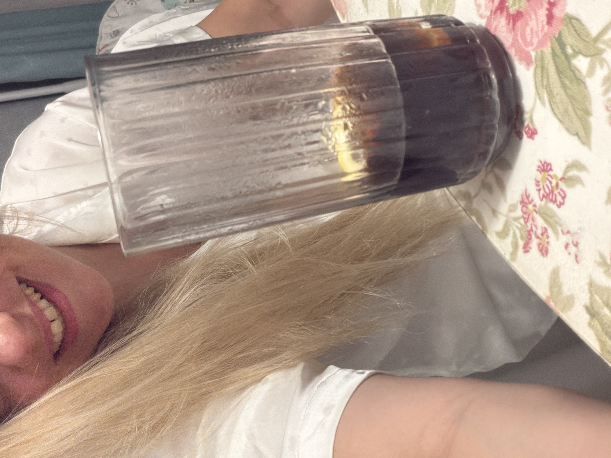 This is an indoor scene occurring in Votkinsk, Russia. The primary subject is a light-skinned individual, likely female, positioned on the left, partially obscured by the foreground. She has long, straight blonde hair extending across her shoulder and chest. Her mouth is open in a smile, revealing her upper teeth which exhibit a yellowish hue and a visible interdental space between the central incisors. She wears a white, short-sleeved garment with a subtle, light-colored embossed pattern, possibly of hearts. In the foreground, centered right, a tall, ridged glass holds a dark brown liquid, suggesting a beverage like cola, with several ice cubes and a visible lemon wedge. The glass rests on a white tablecloth adorned with a repeating floral pattern featuring prominent pink roses, green leaves, and smaller red-pink flowers. The background on the upper right includes blurred light blue-green fabric.
