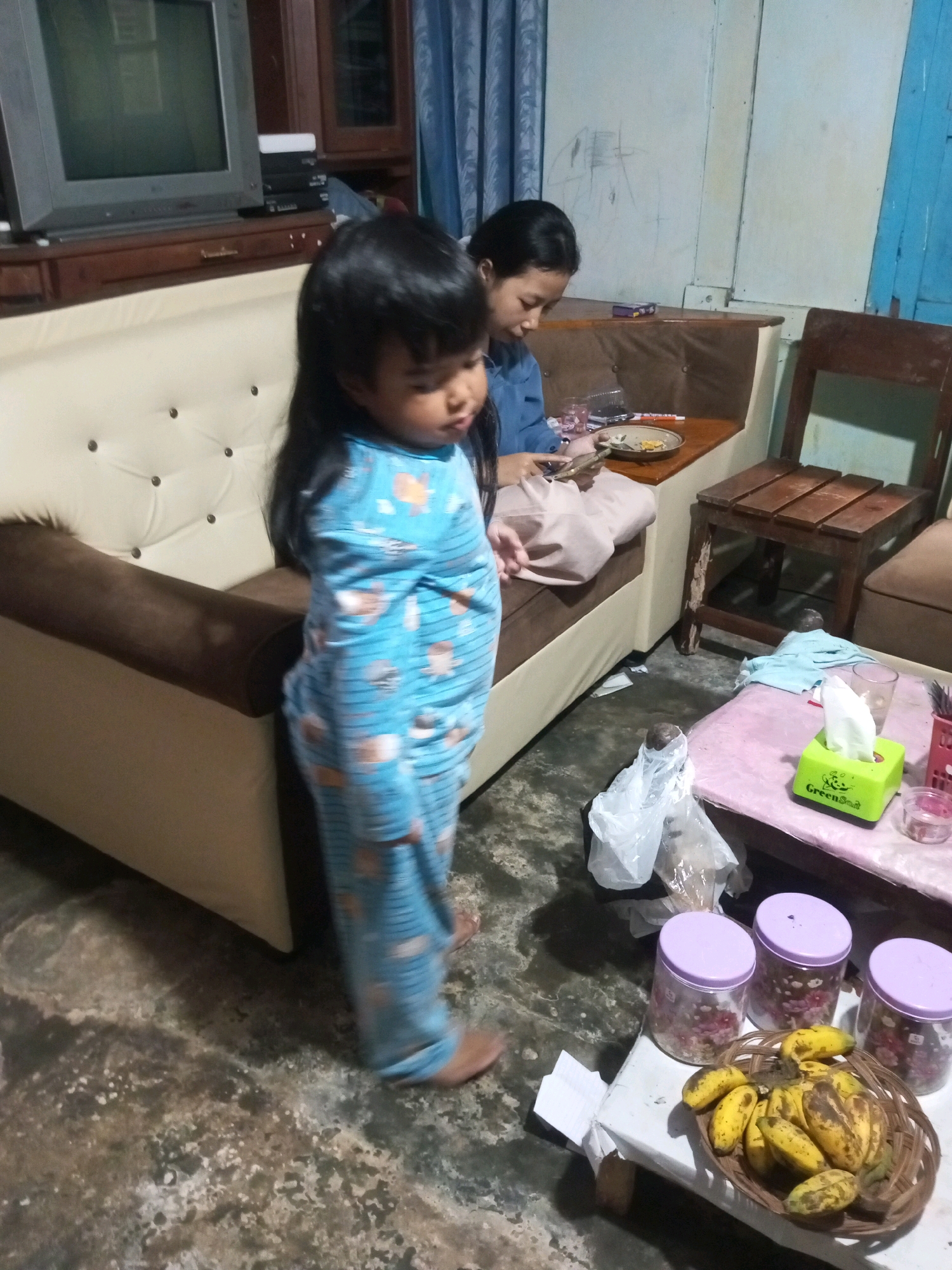 In a dimly lit indoor setting that appears to be a living room in Andongsili, Indonesia, a young child wearing blue pajamas patterned with cartoon animals stands near a sofa. The child's back is partially turned to the camera, and they are looking downwards. To the right of the child, a young woman wearing a blue shirt is seated on the sofa, looking at her phone. In the background, a television set is visible on a wooden cabinet, and a window with blue blinds is partially open. In the foreground, a low table holds a basket of bananas, three jars with purple lids, a tissue box, a glass, and some plastic bags. The floor is concrete and appears worn.