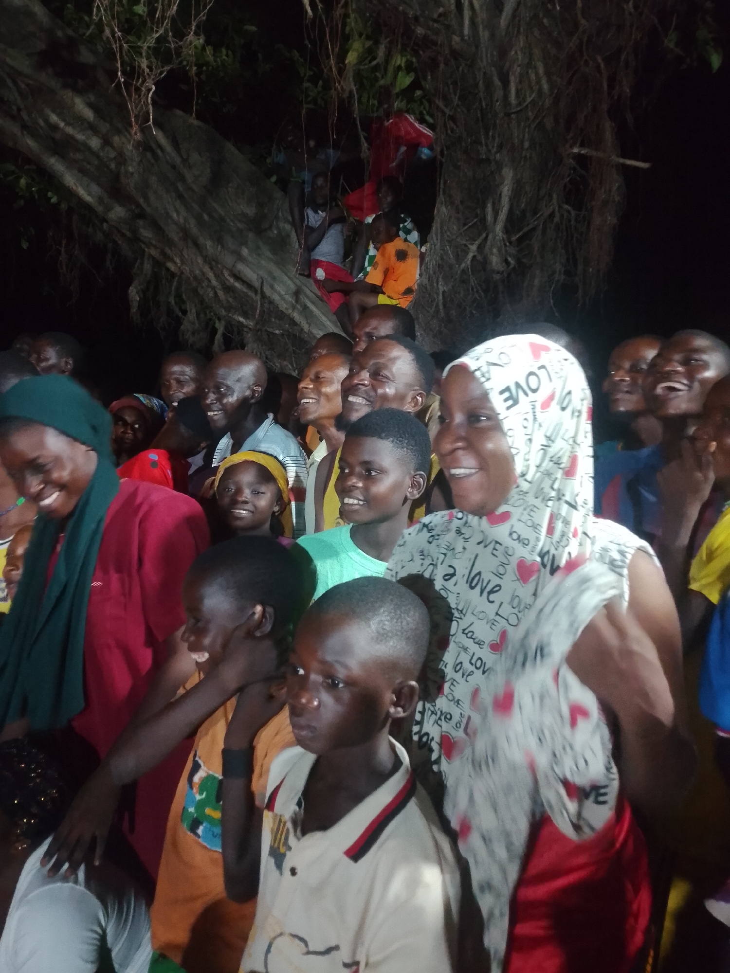 A group of people are gathered outdoors at night, with a large tree in the background. The group appears to be diverse in age, with both adults and children present. Many of the individuals are smiling and looking towards the camera. The lighting suggests it is dark, with artificial lights illuminating the scene. The presence of the large tree and the outdoor setting indicate a natural or semi-natural environment, possibly a park or a village gathering space. Some individuals are wearing headscarves and casual clothing, suggesting a relaxed and informal occasion.