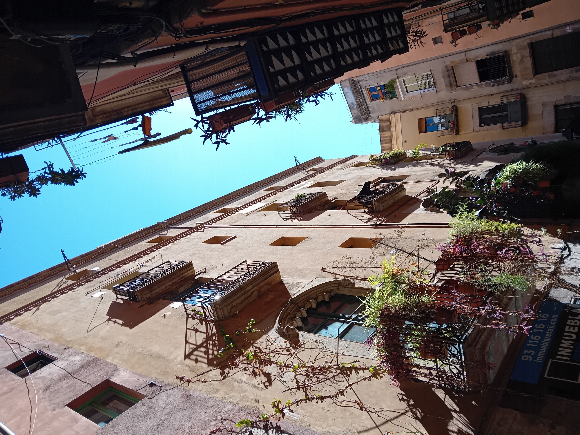 The image is taken from a low angle, looking up at the buildings in a narrow street in Barcelona, Spain. The sky is visible between the rooftops, and is a clear blue color. The buildings on either side of the street are old, with weathered facades and a variety of architectural details. There are multiple windows and balconies, some of which are adorned with plants and flowers. There are also several signs and advertisements visible on the buildings. The overall impression is one of a vibrant, historic city with a unique atmosphere.