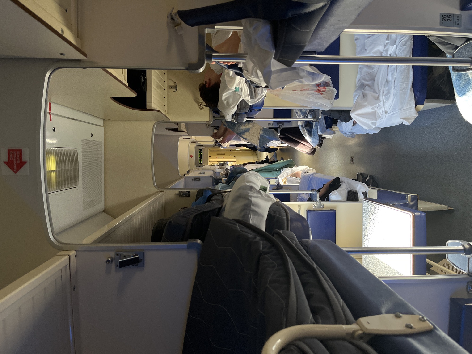 The image depicts the interior of a passenger train. People are lying in bunk beds, covered with sheets and blankets. Bags and personal belongings are scattered around the sleeping compartments. The train appears to be in transit. The location, Dzyakino, Russia, cannot be confirmed from the image.