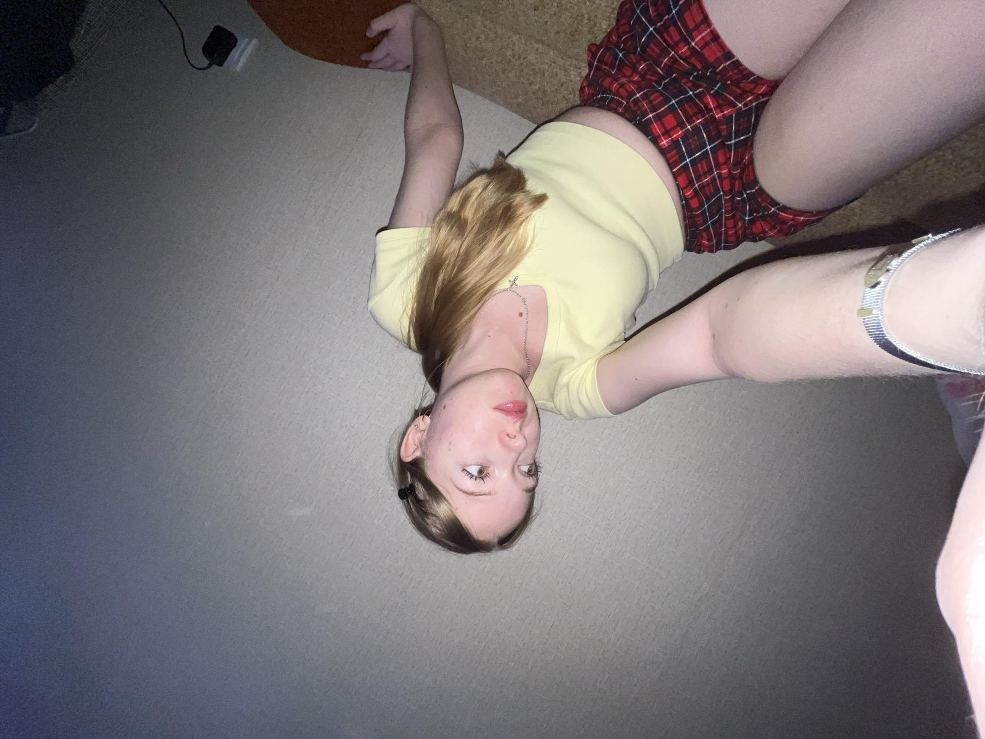 An indoor, overhead shot captures a young person, likely female, lying on the floor and a piece of furniture, with the image rotated so her head is towards the bottom of the frame. She has fair skin, long light brown hair parted to the side, and is looking directly towards the viewer with a neutral to slightly pensive expression, her lips slightly parted. She is wearing a light yellow, short-sleeved top that reveals a small section of her midriff, and a red and black plaid skirt. A silver-toned necklace with a cross pendant hangs around her neck, and a silver-colored bracelet is visible on her right wrist. Her hair is pulled back on one side with a dark hair clip.
She appears to be in a relaxed pose, with her arms outstretched to her sides and her legs bent, resting partly on a light-colored carpeted floor and partly on a brown, upholstered surface, possibly a couch or chair. The background features the same light-colored carpet, showing some texture. In the upper left corner, a black power adapter or charger connected by a cable is visible on the floor. The lighting in the scene is dim and ambient, suggesting an indoor setting without strong direct light, possibly evening. No discernible text is visible anywhere in the image. The scene takes place in Chaikovsky, Russia.