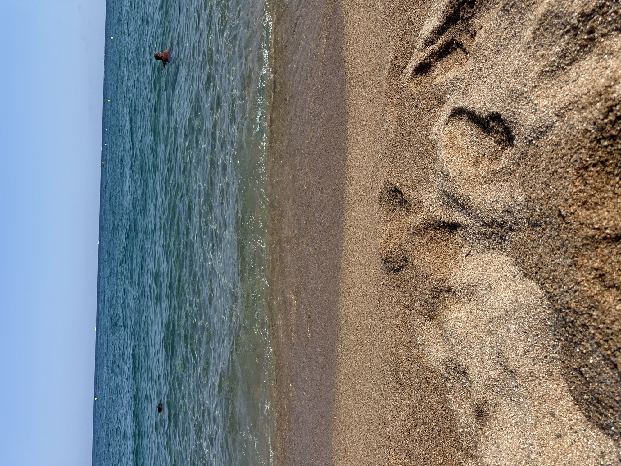 The image captures a view of the Mediterranean Sea from the shore in Blanes, Spain. The foreground is dominated by wet, sandy beach with visible footprints, suggesting recent human activity. In the middle ground, the clear, shallow water laps gently at the shore, revealing the sandy seabed beneath. Further out, the deeper ocean exhibits a turquoise hue, with a few yellow buoys and distant boats dotting the horizon. Two individuals are present in the water; one is partially submerged and appears to be swimming, while another object, possibly a person or an animal, is visible as a dark shape further to the left. The sky above is a clear, cloudless blue.