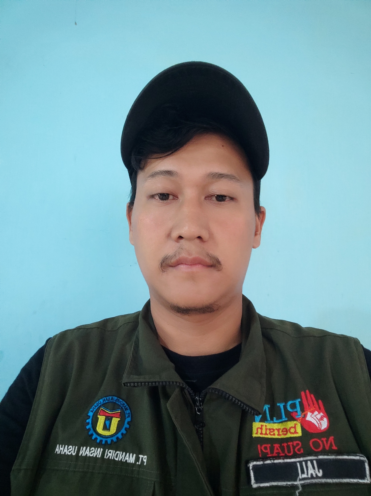 A man wearing a dark green vest over a black shirt and a black baseball cap poses for a selfie in front of a light blue wall. The man has short dark hair, a mustache, and a goatee. The vest has several patches and embroidery. On the left breast is a circular patch with a blue and yellow logo and text. Below this patch, text is embroidered in white: "AHASU ИAS ИI ЯIDNAM Tg". On the right breast, there are two embroidered elements. The top element is colorful, featuring red and blue text that reads "PLN" and a red hand symbol. Below this, red text reads "diered 19AUS2 ON". A rectangular black patch with white text saying "JALL" is attached below the right breast. The background is a plain, light blue wall. The lighting is direct and appears to be from a flash. The scene is indoors, likely in a room or hallway in Gebang, Indonesia.