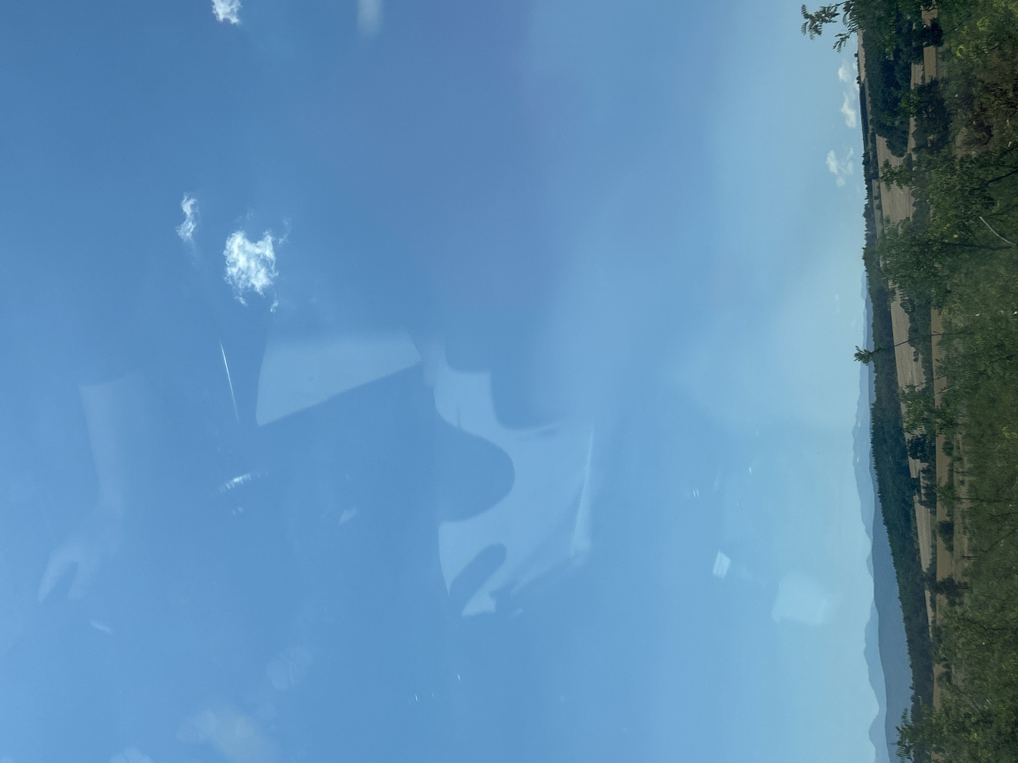 The image shows a bright blue sky with scattered white clouds. There are reflections on the surface of the sky, suggesting it might be viewed through a window. In the distance, there are rolling hills and mountains, covered with a mix of trees and fields. The terrain appears to be rural and somewhat arid. The colors are muted, with shades of green, brown, and blue dominating the scene. There are no people or animals visible in the image. The overall impression is one of a serene, natural landscape on a clear day. The exact location in Vakarel, Bulgaria, cannot be determined from the image alone, but the scenery is consistent with a rural setting in that region.