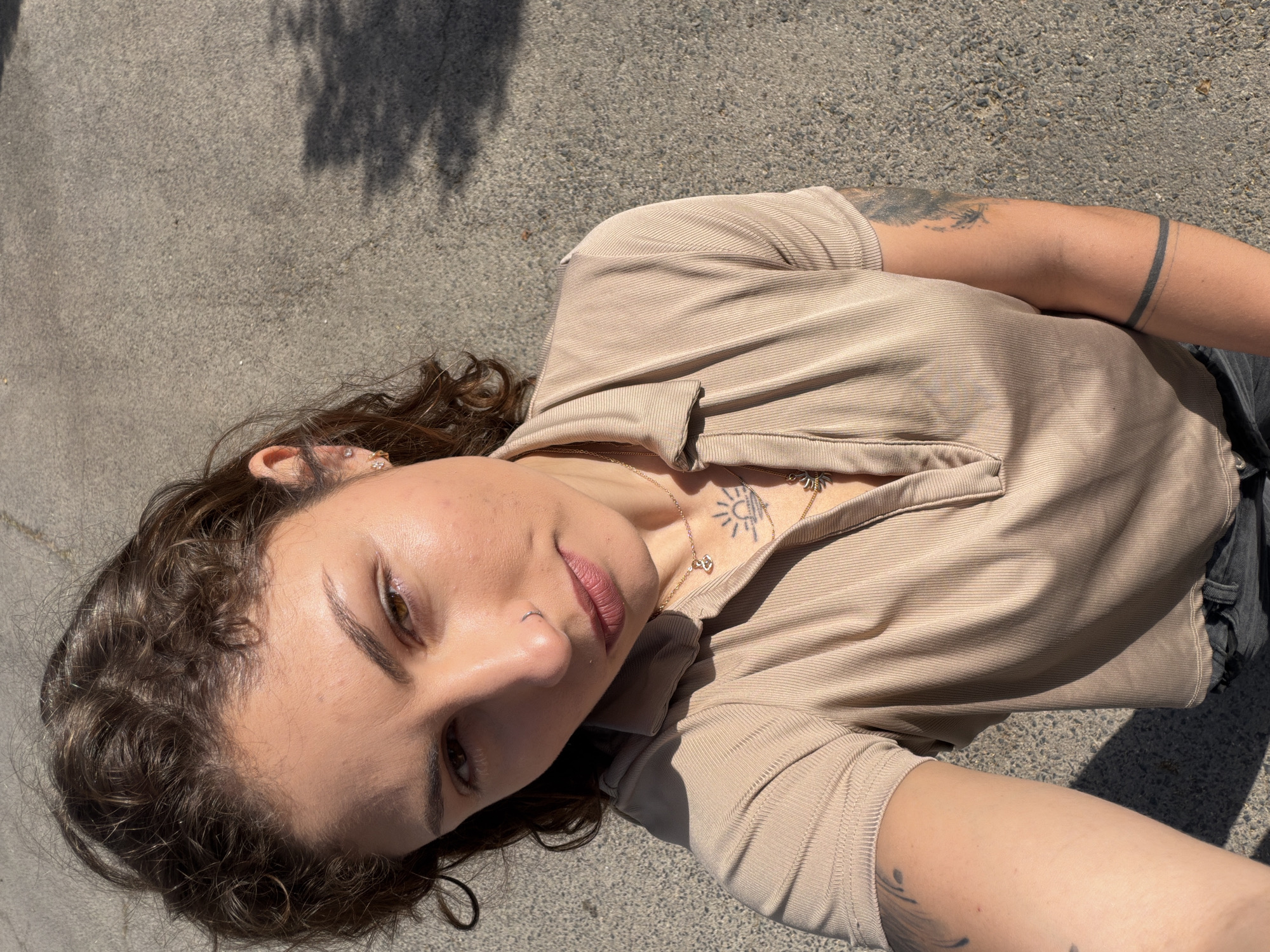 A woman lies on a grey asphalt surface in Kiryat Bialik, Israel. She faces the camera and has brown, curly hair, light skin, and brown eyes. She wears a beige, short-sleeved, button-down shirt. A gold chain necklace is visible. The woman has tattoos on her chest and both arms. The background is a plain asphalt surface, partially in shadow.