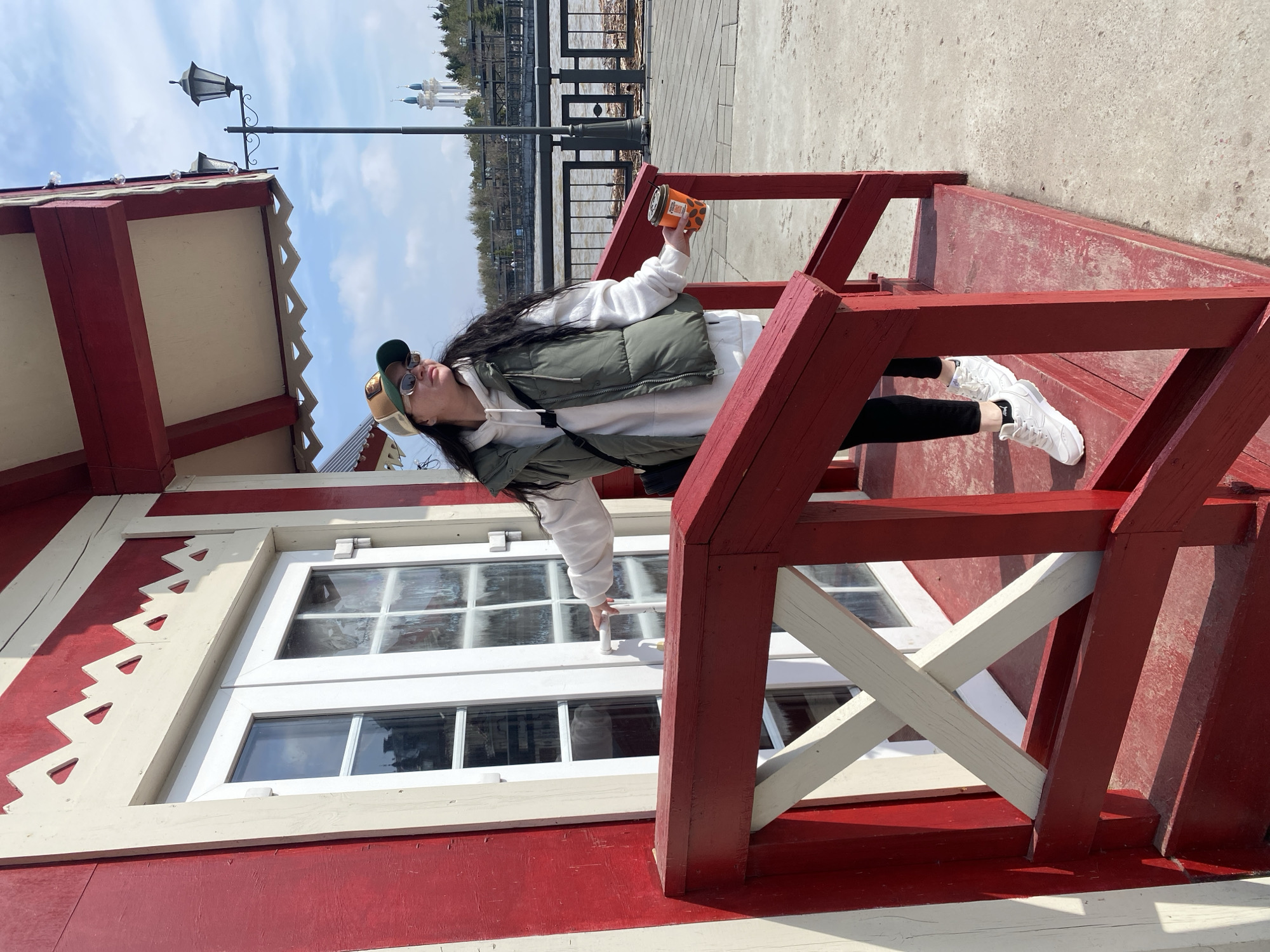 A young woman stands on a red-painted wooden balcony with white accents, holding a disposable coffee cup and looking toward the background, in Kazan, Russia. She wears a white hoodie, an olive-green puffer vest, black leggings, and white sneakers, as well as a cap and sunglasses. The balcony is part of a building with white-framed windows. Beyond the balcony, the view includes a body of water, a distant cityscape with notable buildings, and a lamppost. The sky is partly cloudy.