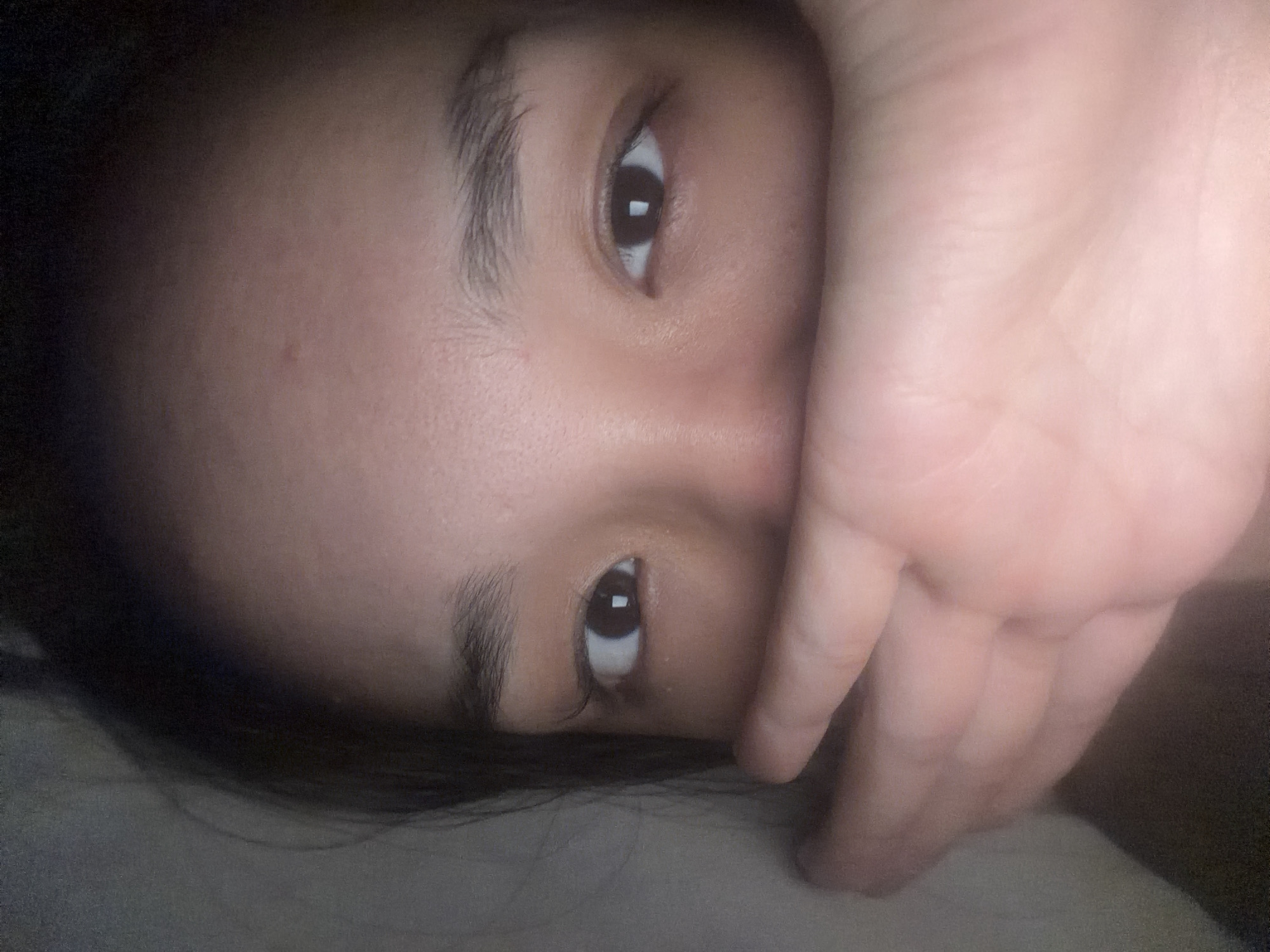 The image is a close-up, low-angle shot of a person's face partially obscured by their own hand. The person appears to be lying down, as indicated by the soft, textured surface beneath them, possibly a pillow or blanket. The lighting is dim and warm, creating soft shadows across the face. The visible eye is dark and open, with a reflection visible in the pupil. The hand is positioned to cover the mouth and nose area of the face, with visible skin texture and creases. The person's hair is dark and frames the visible part of their face. There is no discernible background that would indicate a specific location within Sinyavino, Russia, or any other city. The image focuses solely on the intimate details of the person's face and hand.