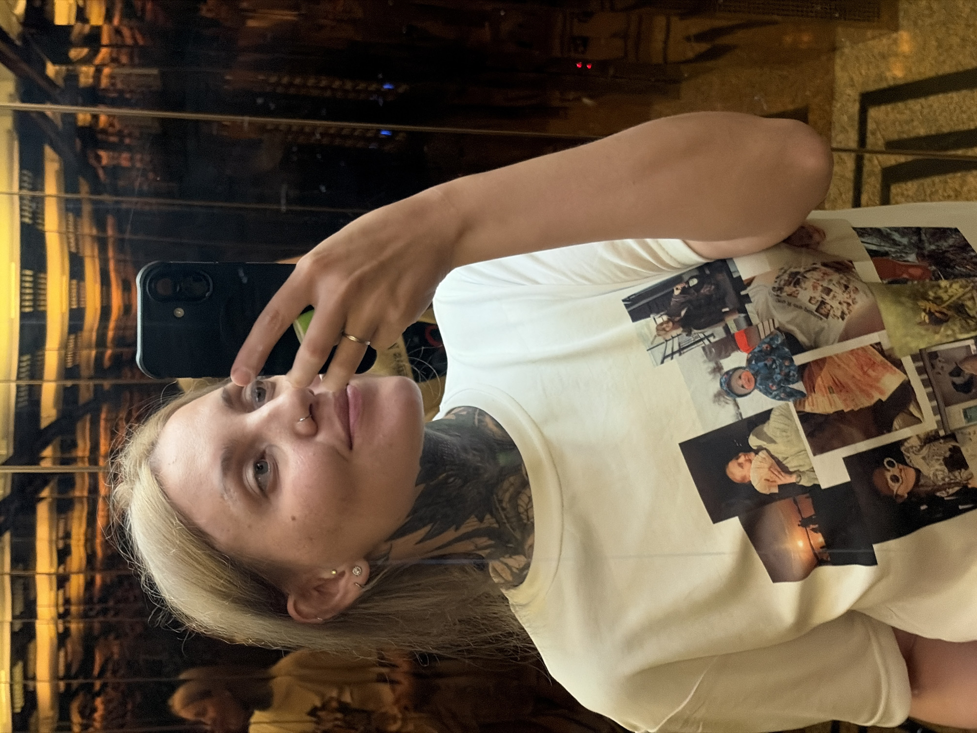 A woman with blond hair and tattoos is taking a selfie in front of a reflective gold-colored wall. She is holding a black smartphone with a camera. The reflection shows a slightly blurred and distorted environment, possibly an interior with decorative elements. The woman is wearing a white t-shirt with a collage of printed images on the front. She has multiple piercings on her ear and a ring on her finger. The location appears to be indoors. There is no indication that this image was taken in Da Nang, Vietnam.