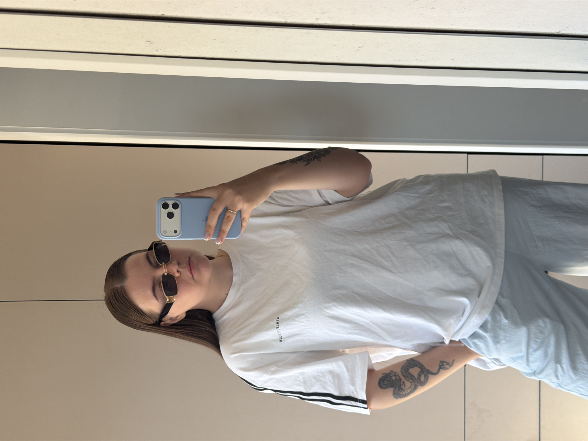 A young woman is taking a mirror selfie with a light blue iPhone. She is wearing sunglasses, a white t-shirt with black stripes on the sleeves, and light blue pants. She has a tattoo on her forearm. The location appears to be indoors, possibly a dressing room or bathroom. The mirror is framed by white trim. The city of Chaikovsky, Russia cannot be confirmed.