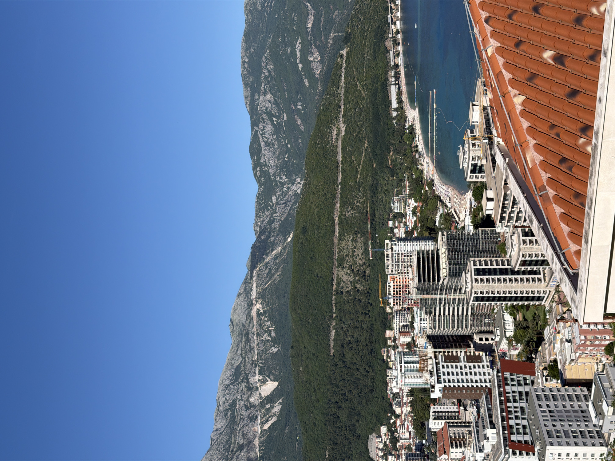 The image depicts a partial view of Budva, Montenegro, from an elevated perspective. The Adriatic Sea is visible along the coastline, with a crowded beach and a long swimming area defined by barriers. Buildings, mostly high-rise apartments and hotels, dominate the foreground. The scene is bordered by a green mountain range to the left and a glimpse of terracotta rooftops in the extreme right corner. The sky is a clear, bright blue. Construction cranes are visible among the buildings, indicating ongoing development. The overall impression is of a popular coastal resort city nestled between mountains and the sea.