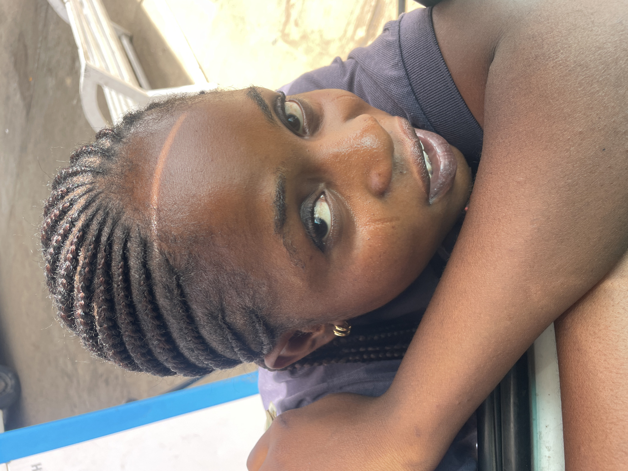 The close-up, eye-level shot focuses on a young Black female outdoors, with her face turned towards the camera and her head resting on her arm. She has her hair braided in thin cornrows. A faint orange stripe of light crosses her forehead. Her lips are slightly parted, revealing her teeth. She is wearing a dark purple collared shirt, and a small gold hoop earring is visible in her left ear. Her right arm is bent, supporting her head as she leans forward. The background shows a blurred outdoor setting with a white bench and a portion of a blue and white structure. The scene is illuminated by natural sunlight, casting shadows and highlighting the texture of her skin.