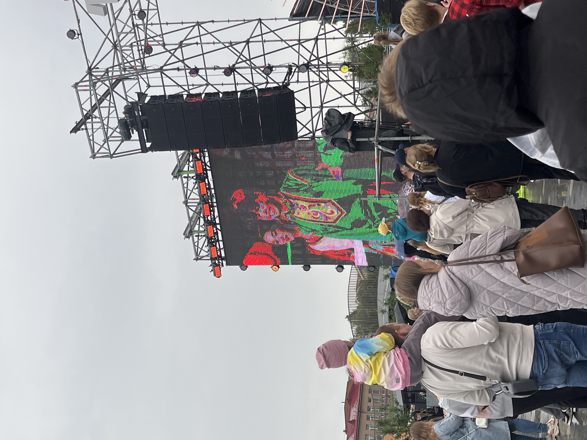 This is a wide-angle, eye-level shot of an outdoor event, likely a concert or festival, under an overcast sky. The primary focus is a large LED screen displaying a video of two performers dressed in vibrant, traditional-looking costumes. The male performer on the right wears an elaborate green and gold outfit. In the foreground, a diverse crowd of people is gathered, facing the screen. Some individuals are carrying children on their shoulders, including a young child in a colorful jacket and a pink hat perched on their father's shoulders. Others are walking or standing, watching the performance. A large sound system and scaffolding are visible to the right of the screen. In the background, buildings and trees suggest an urban or park setting. The ground appears wet, indicating recent rain. This event is taking place in the city of Perm, Russia, specifically at the Perm city exhibition center.