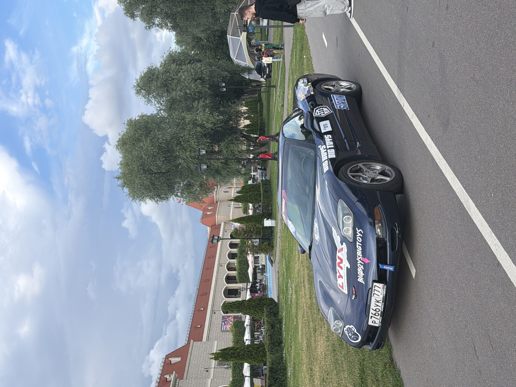 An outdoor scene shows a dark blue Chevrolet Corvette parked on an asphalt road next to a grassy area. The car has several stickers on its hood and sides, including "LYNX", "BIG BOYS BIG TOYS", "BT5", "110", and "SIRON". A license plate that reads "P766YK 777" is visible on the front of the car. A white line marks the edge of the road. To the right of the car, a man wearing a black hoodie and light-colored pants stands facing the car. In the background, a large building with red roofs and arches is visible, surrounded by trees and manicured bushes. Further in the background, there are people walking and a small fountain area. The sky is partly cloudy.
