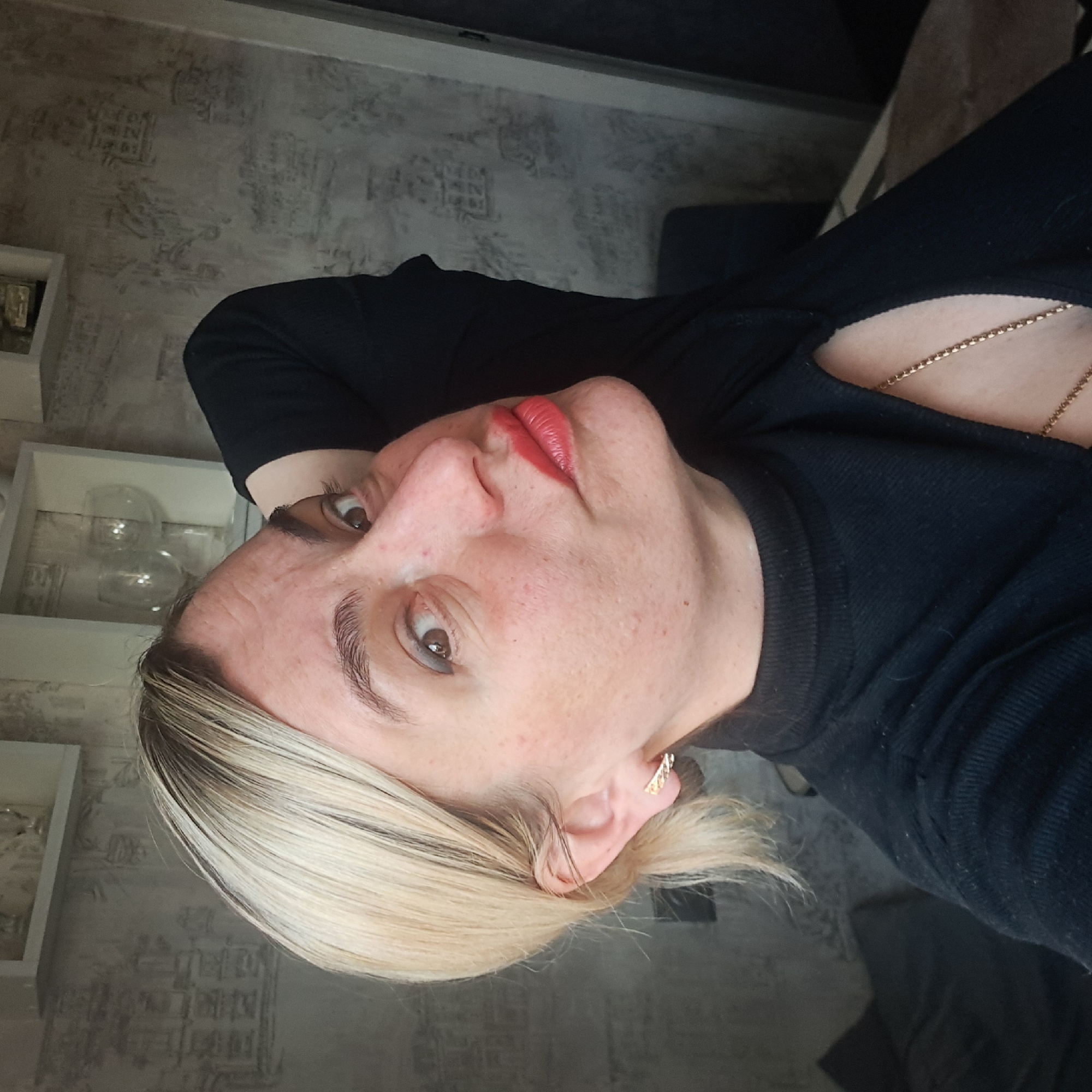 A medium-shot photograph, taken indoors, depicts a woman in a black top. She is wearing red lipstick and a gold necklace and earring. She has short, blonde hair. The background consists of a patterned wall, a display shelf, and possibly a chair or piece of furniture. There is no specific information about the city of Elektrostal, Russia.