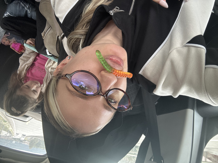 The image displays two females within a moving vehicle, identified as being in Menteşe, Turkey. In the foreground, a female with blonde hair and dark-framed oval eyeglasses is visible, facing forward. An orange and green gummy worm is positioned horizontally between her upper lip and nose. She wears a black jacket featuring beige and white panels. A light brown, zippered shoulder bag is partially visible on her right side. In the background, seated in the rear passenger seat, a young female child with dark hair in a ponytail is observed. She is looking down at an open book or tablet held in her lap, with a doll partially visible beneath it. The external environment, seen through the vehicle's rear window, shows a multi-lane road with distant vehicles, flanked by vegetated hills.