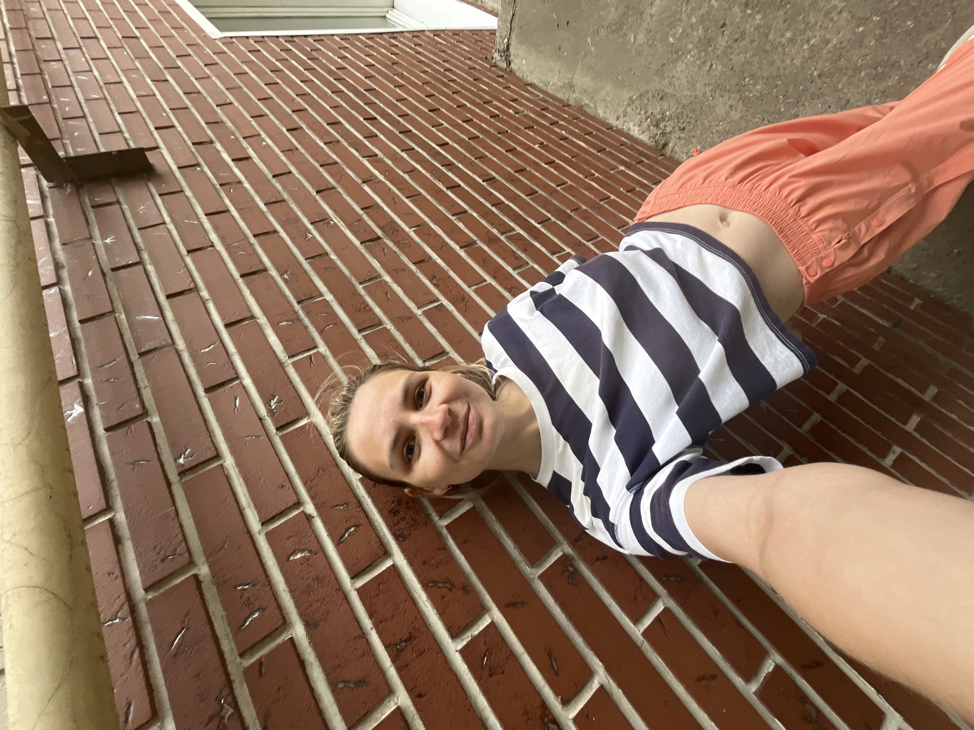 A woman with blonde hair pulled back is lying on a brick wall outdoors, taking a selfie. She is wearing a navy and white striped shirt and peach-colored pants. A small section of her abdomen is visible between the shirt and pants. The brick wall is made of red bricks with white mortar. The background includes a window frame and a section of a concrete wall. The location cannot be determined to be the city of Chaikovsky, Russia based on the visual information.